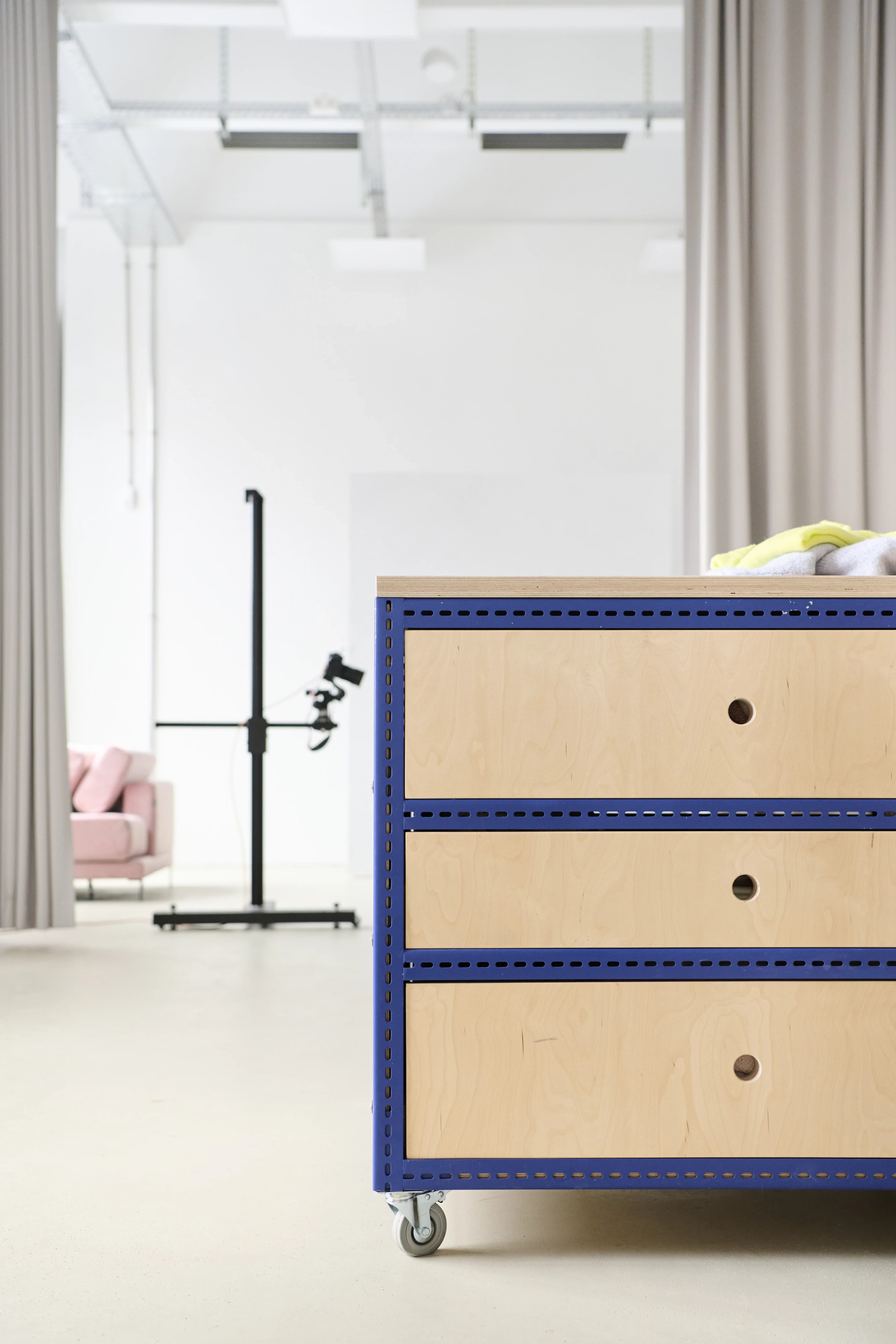 Detail of a mobile birch plywood drawer module with blue perforated steel frame at the FLACONI CONTENT STUDIO, with a photo stand and pink sofa in the background.
