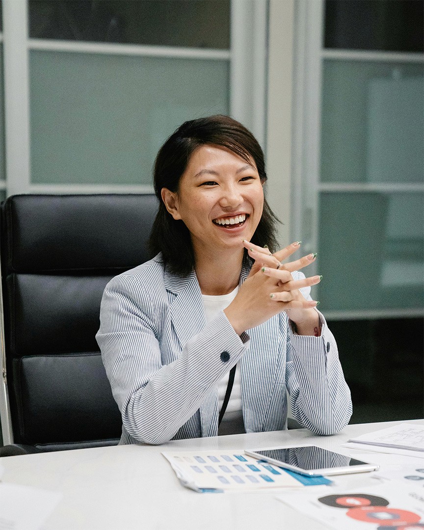 A professional asian woman with her hands clasped in front of her, smiling in an office setting