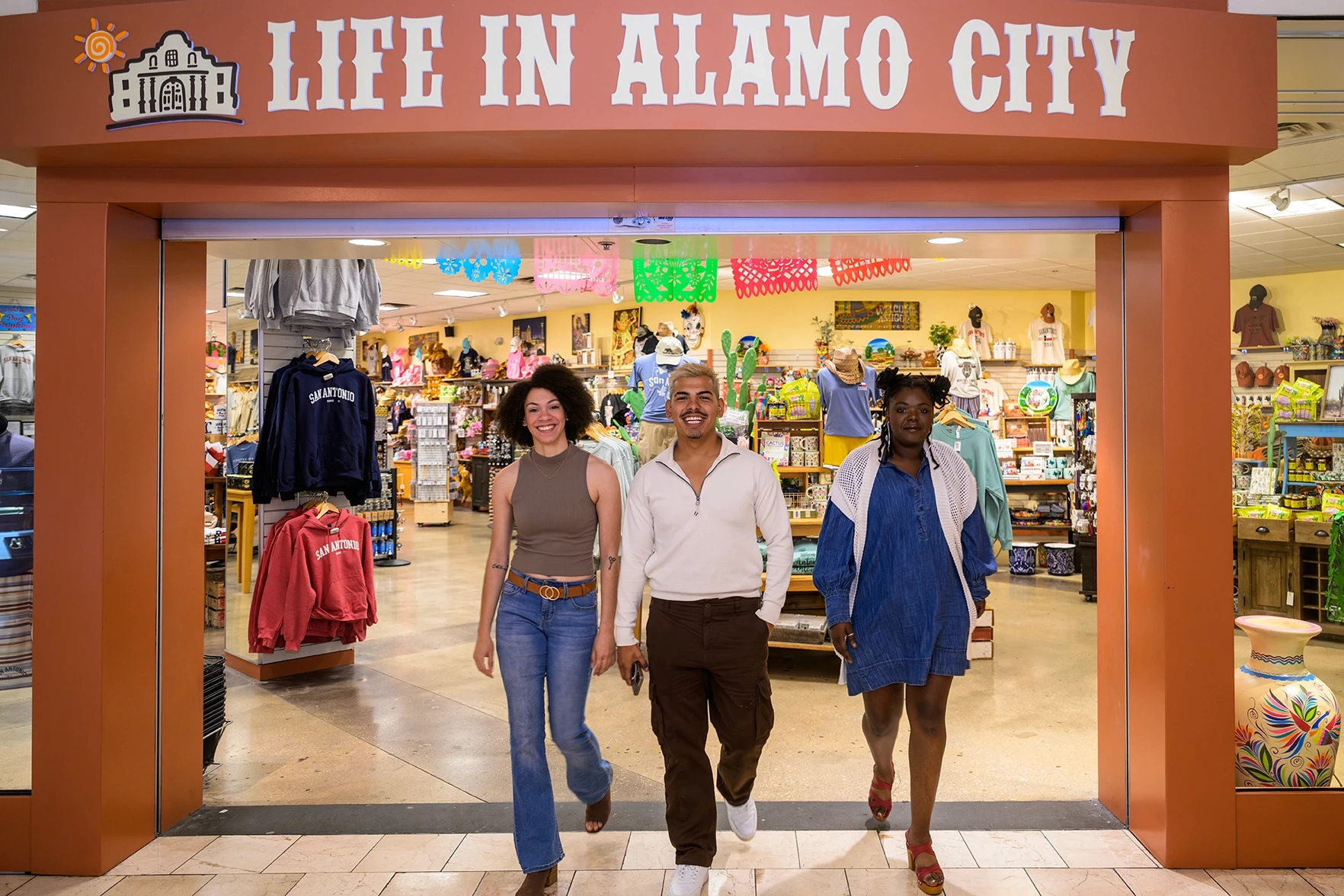 Three shoppers walking out of Life in Alamo City gift shop with colorful merchandise and papel picado decorations visible inside
