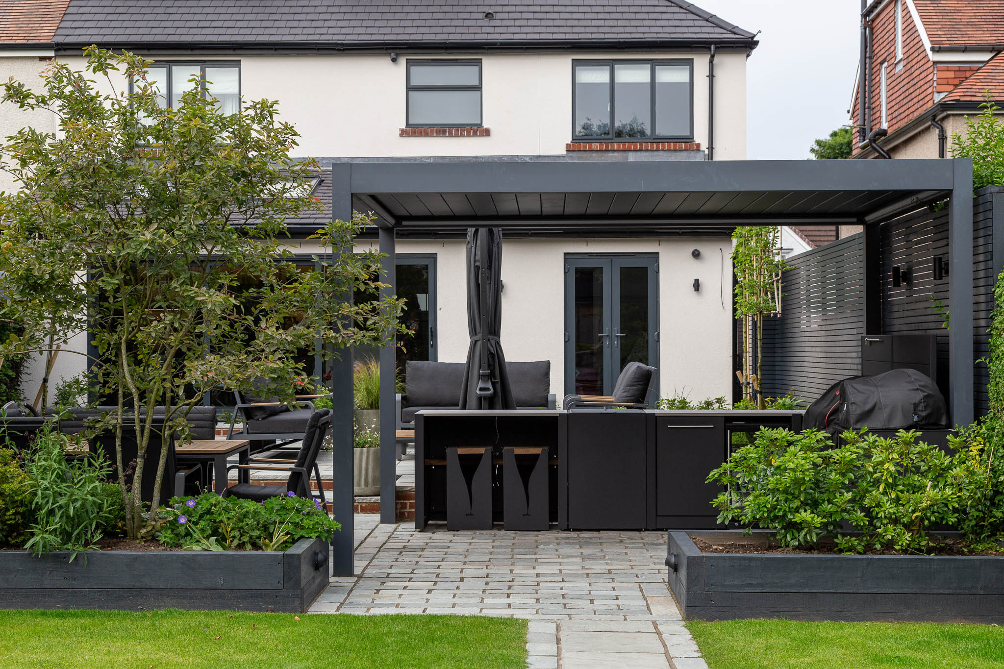 Two black cushioned chairs sit at a wooden table in a lush garden surrounded by vibrant green foliage and trees, creating a serene outdoor space.