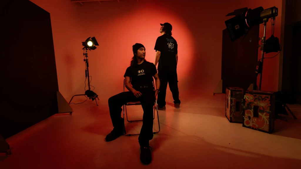 Two men in black streetwear pose in a moody, red-lit studio with professional production gear and spotlights.