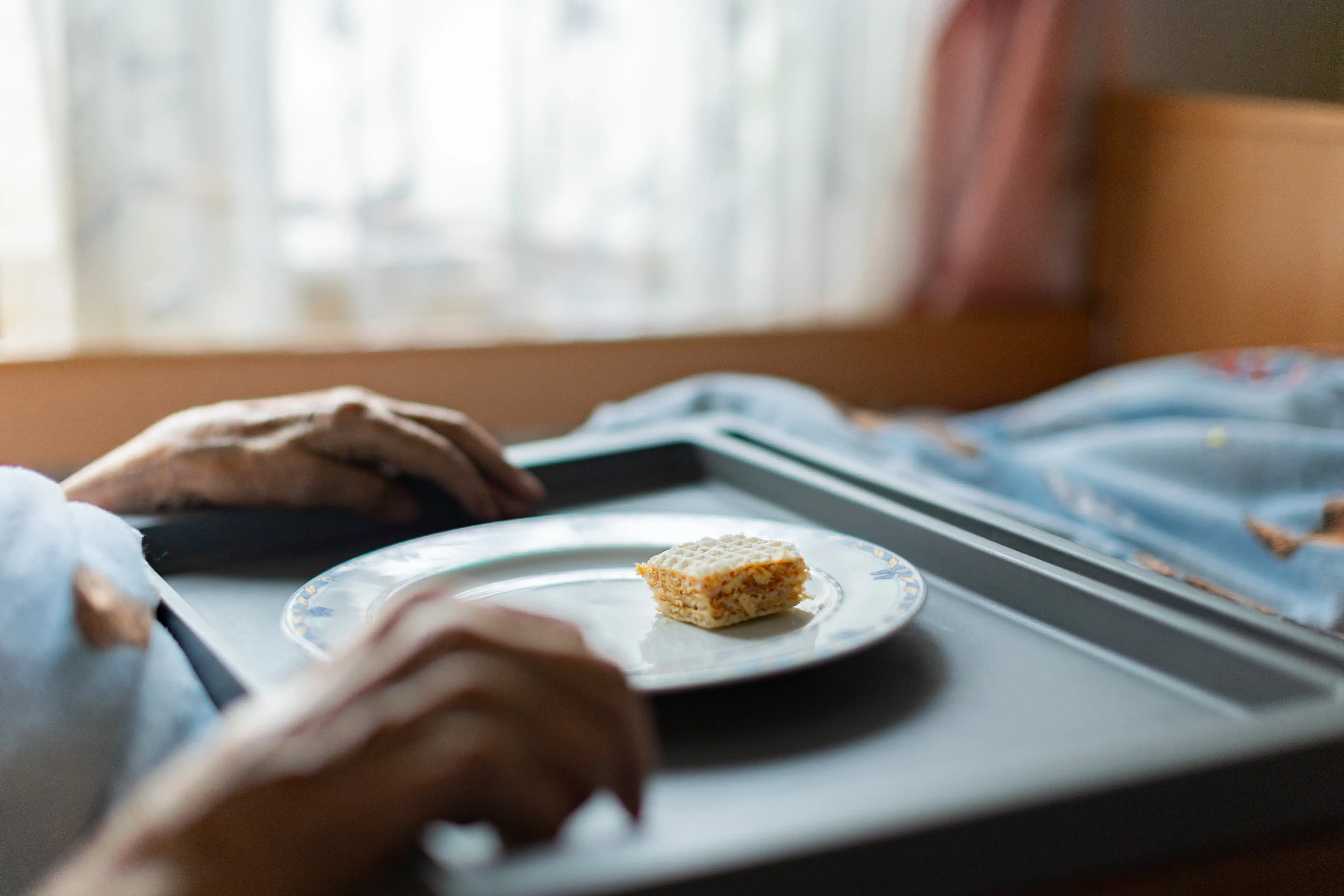 Ręce osoby położone na tacy z talerzem z resztkami jedzenia. W tle okno z firanką oraz niebieska pościel na łóżku.