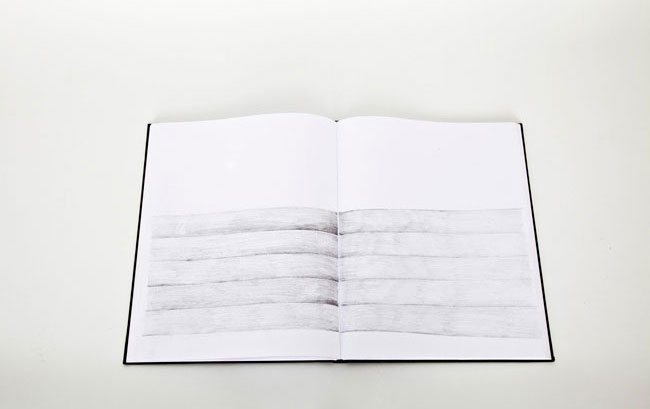 The image shows an open book with a black cover, resting on a white surface. The pages are white and feature a pencil sketch of horizontal lines. The sketch is rendered in shades of gray and black. The lines vary in thickness and create a sense of depth and layering. The sketch is centered on the page, with no text or other objects present.