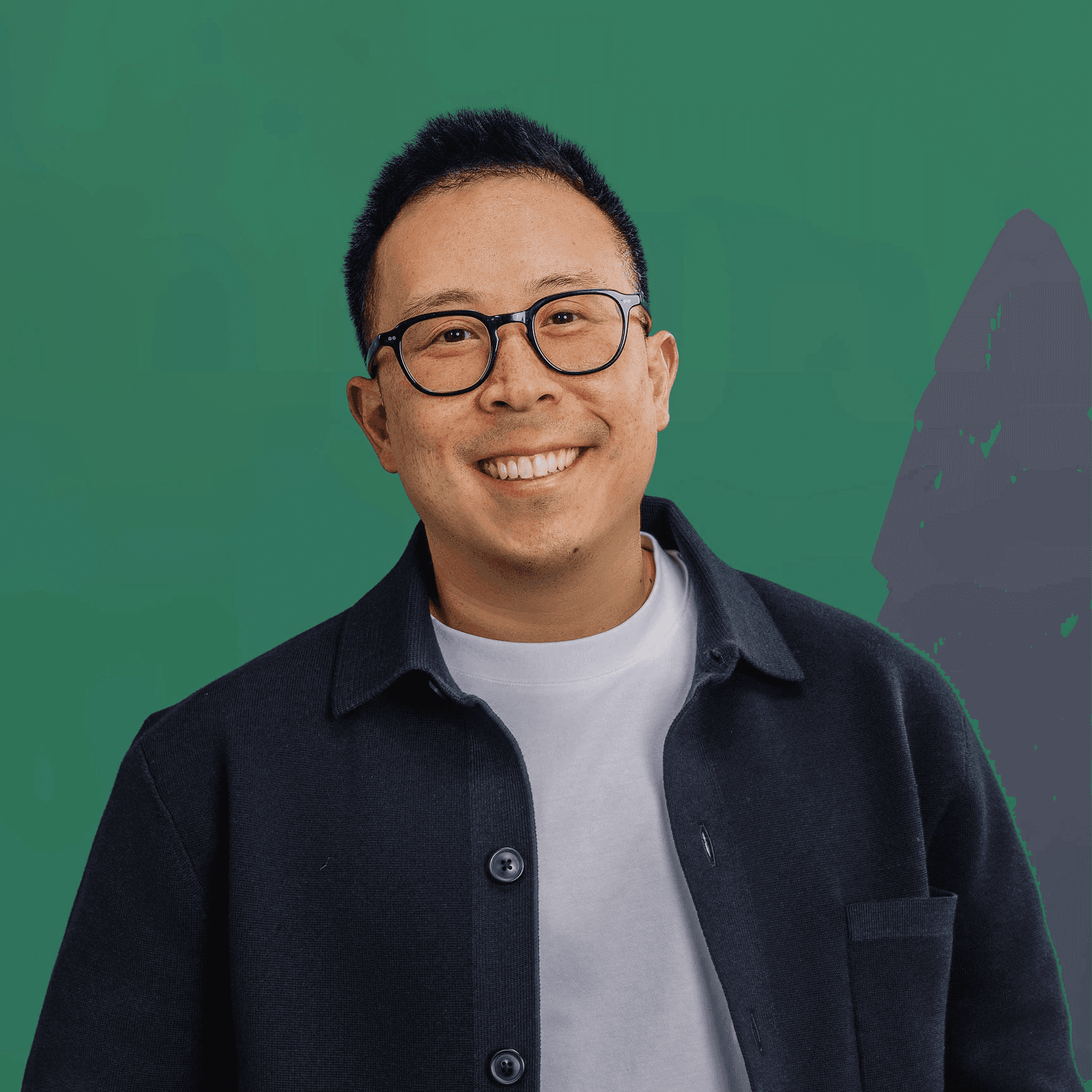 Smiling man wearing glasses and a dark shirt layered over a white tee, standing against a green background.