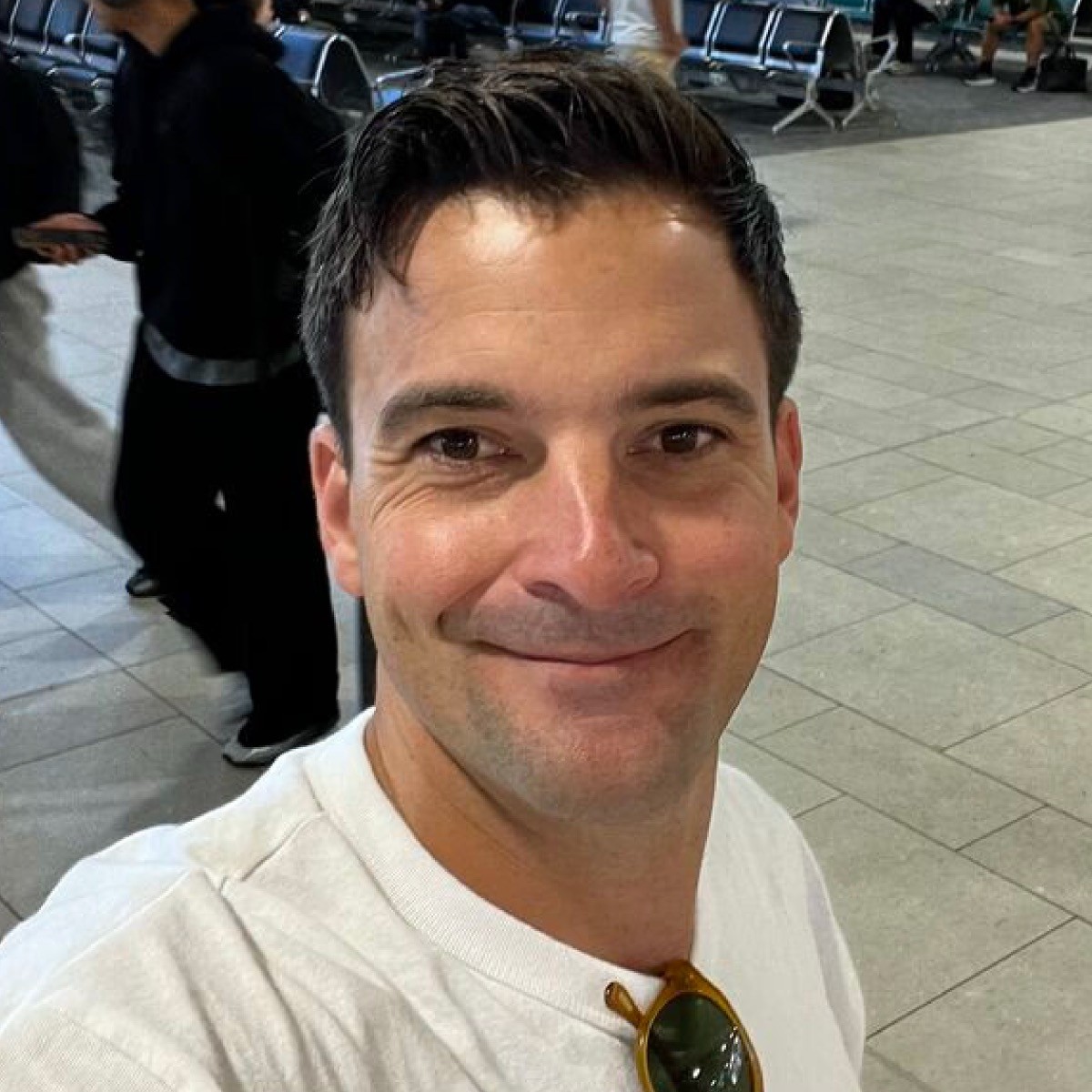 A man wearing glasses, a black hat, and a dark jacket looks confidently at the camera against a light background.