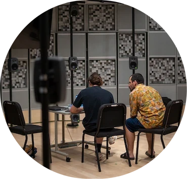 Deux personnes assises de dos devant un ordinateur dans un studio immersif équipé de multiples haut-parleurs suspendus et de panneaux acoustiques.