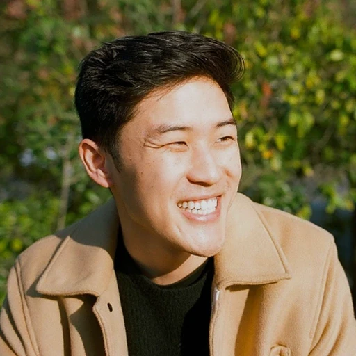 Uma pessoa sorridente, vestindo um casaco bege e uma camisa preta, está ao ar livre diante de uma folhagem verde exuberante, aproveitando um dia ensolarado.