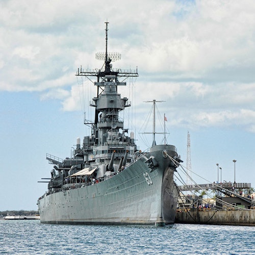 เรือรบสีเทาขนาดใหญ่จอดเทียบท่าที่ท่าเรือ มีเสาอากาศและป้อมปืนที่มองเห็นได้ชัดเจน ท่ามกลางท้องฟ้าที่มีเมฆครึ้ม