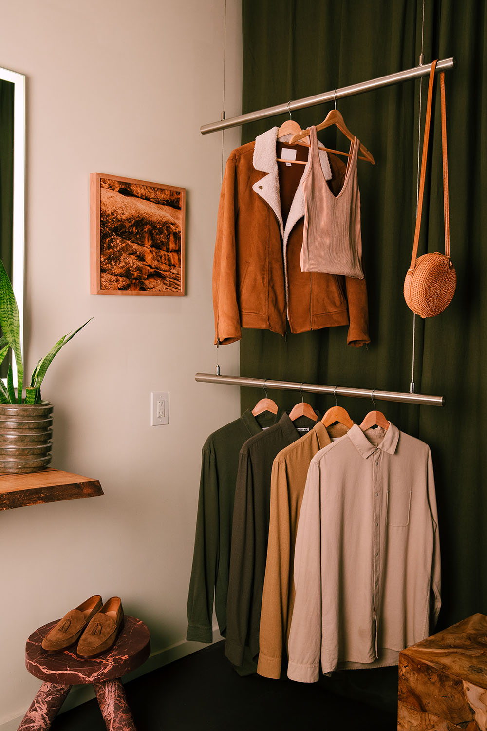 Clothing is artfully hung from a modern clothing rack featured in the greenroom of Particle Studio in Seattle, WA. There are several shirts, a jacket, a tank top, and a wicker handbag. Shoes sit next to the rack on a stone marble stool.