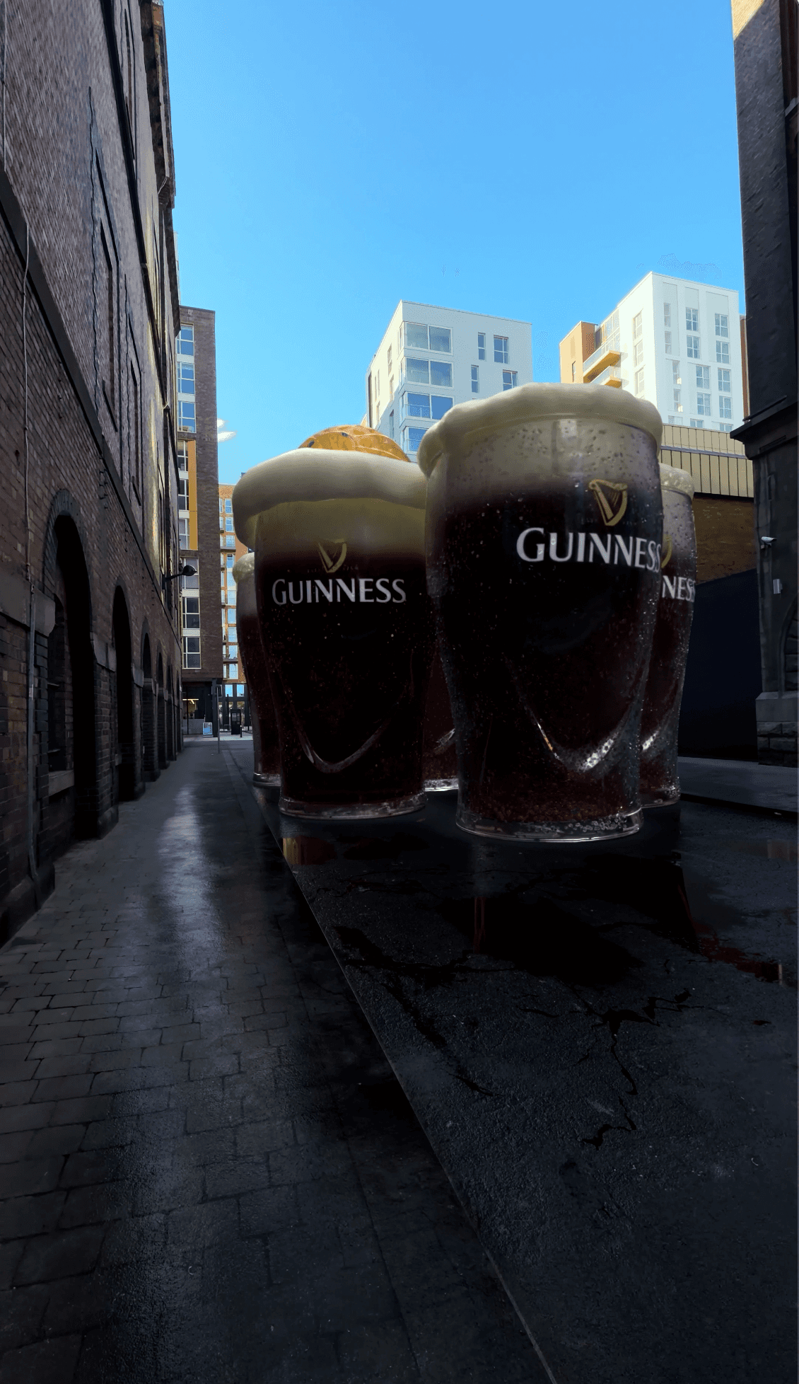 Three giant, hyper-realistic Guinness glasses standing in a city alleyway.