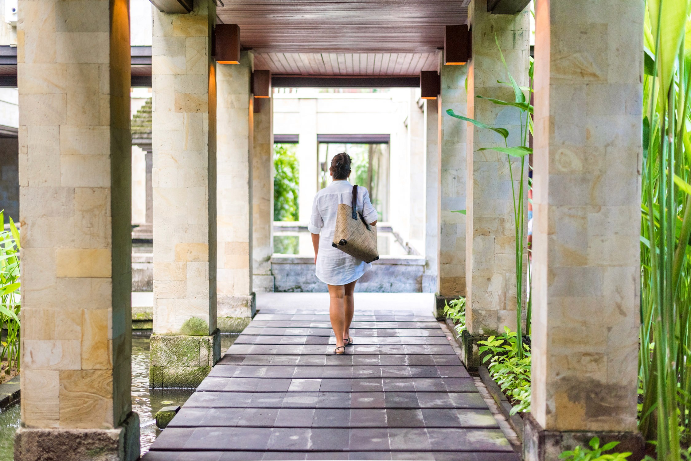 A woman in a white dress and sun hat walks through a stone-pillared corridor toward an open doorway at a resort.