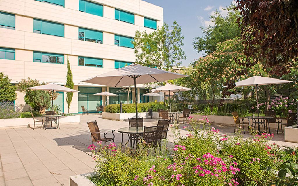 Modern residential building with turquoise balconies, patio area with lounge chairs and umbrella, vibrant flower beds with red and pink blooms, and surrounding greenery
