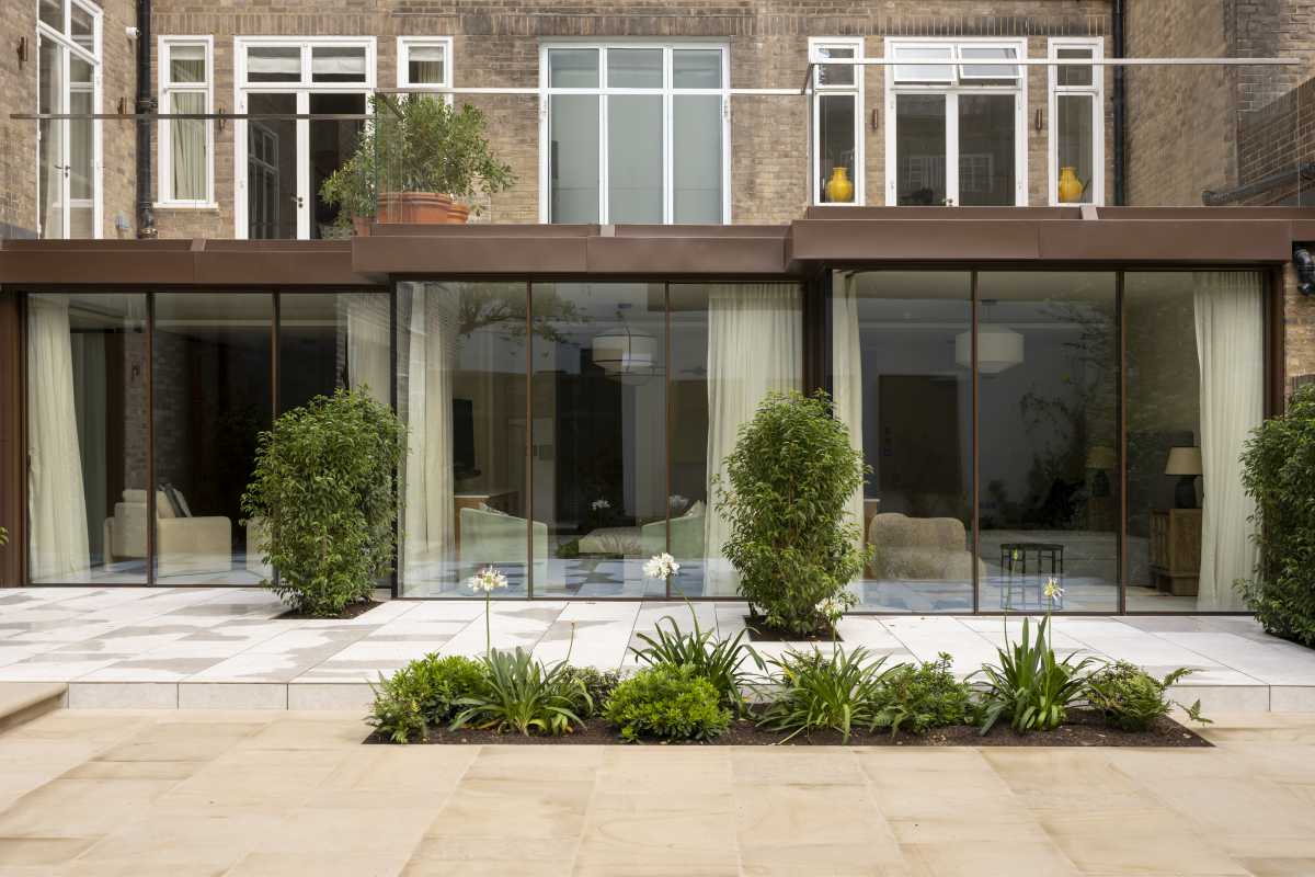 A contemporary full-width glass rear extension on a premium London home, featuring floor-to-ceiling sliding glass doors, a stone patio terrace, and manicured garden planters.