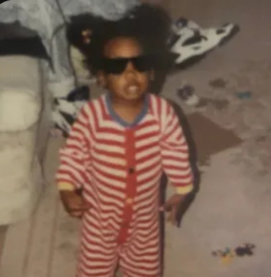 A young child wearing sunglasses and a striped pajamas stands confidently on a carpeted floor.