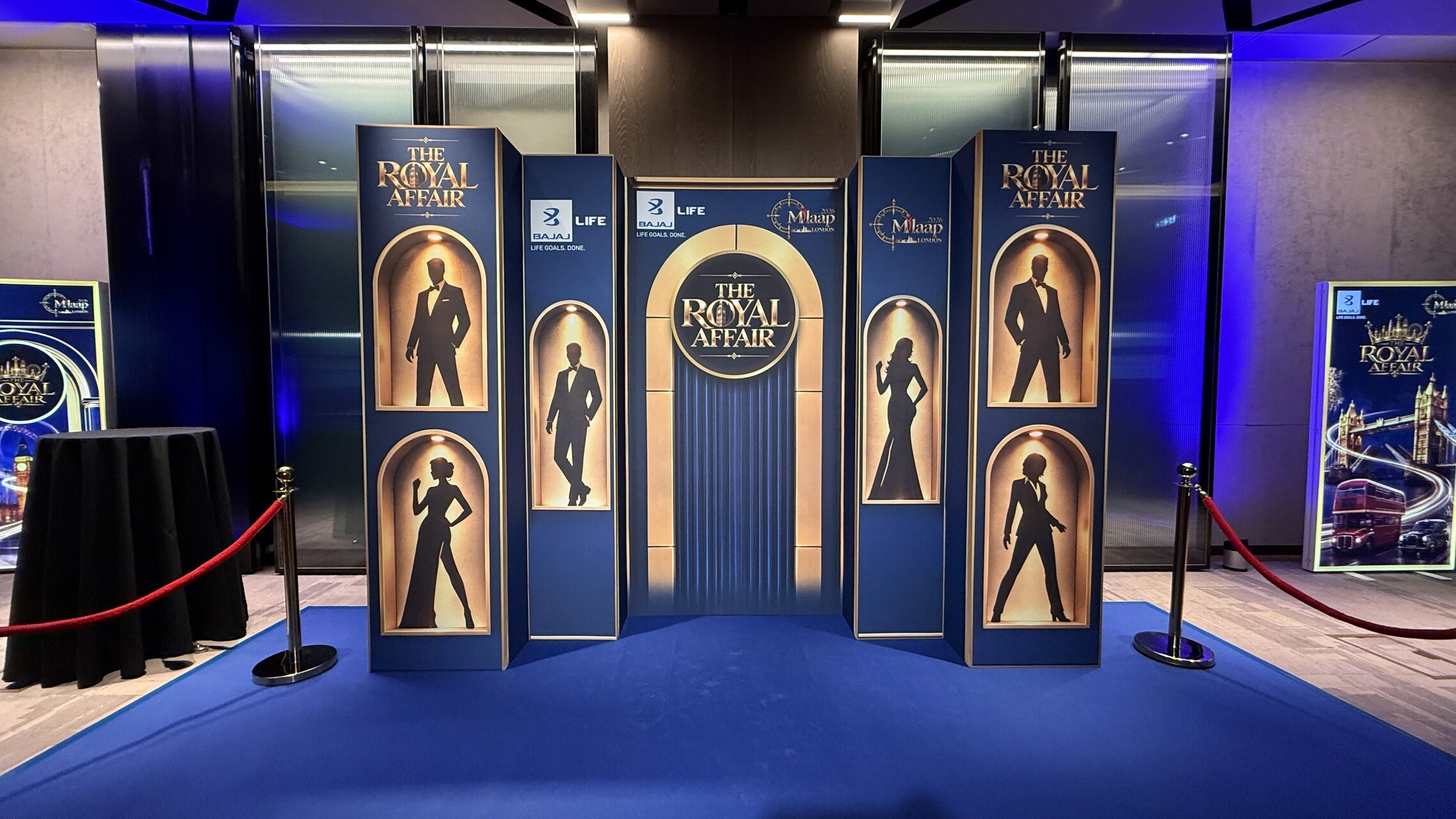 Blue and gold silhouette photo backdrop with a central arch, Bajaj Life branding and red rope barriers.