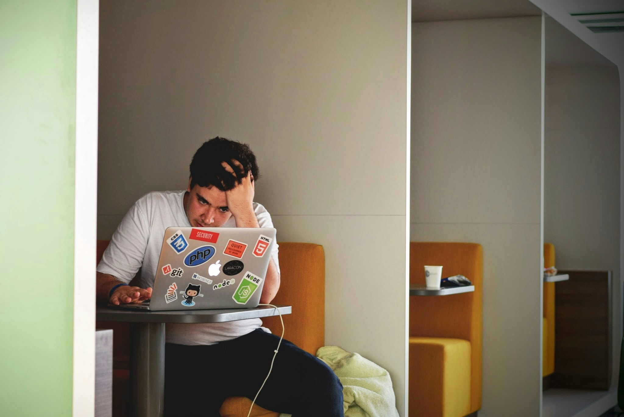 A man in a library cubicle looks stressed. He has his hand on his head and looks down at a sticker-covered laptop.