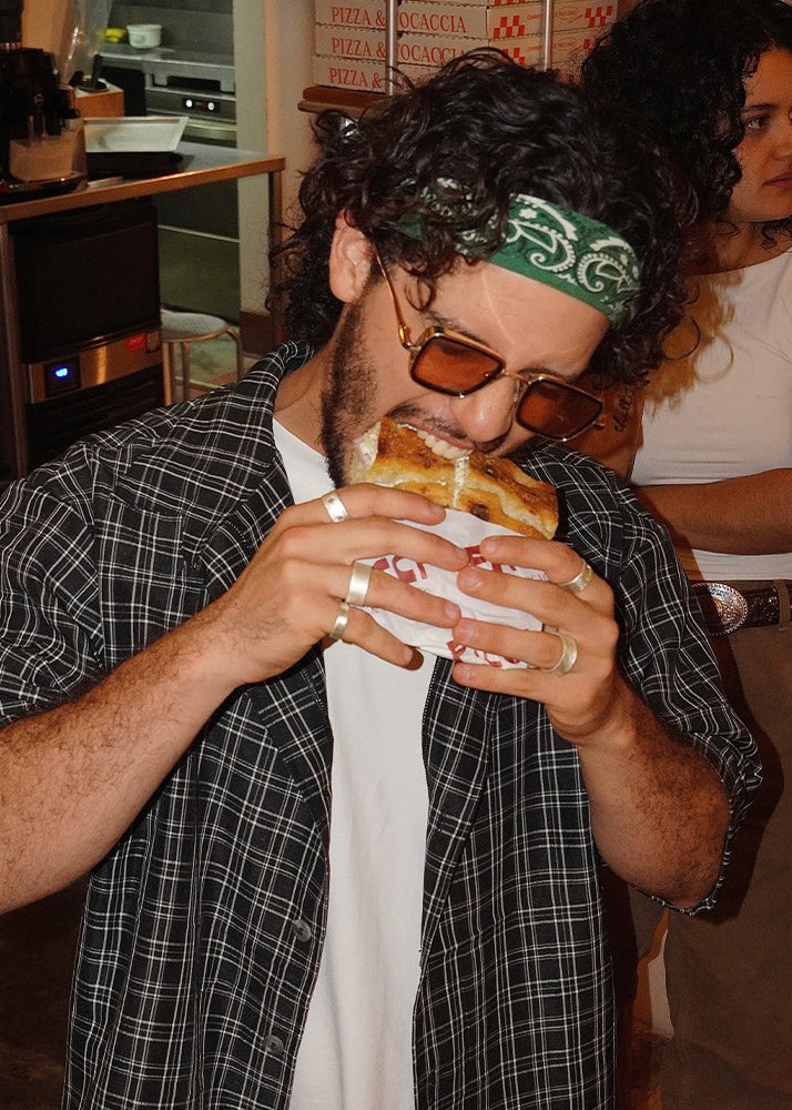 Man eating a sandwich wearing a flannel shirt with silver rings on both hands on a pizza restaurant setting standing up