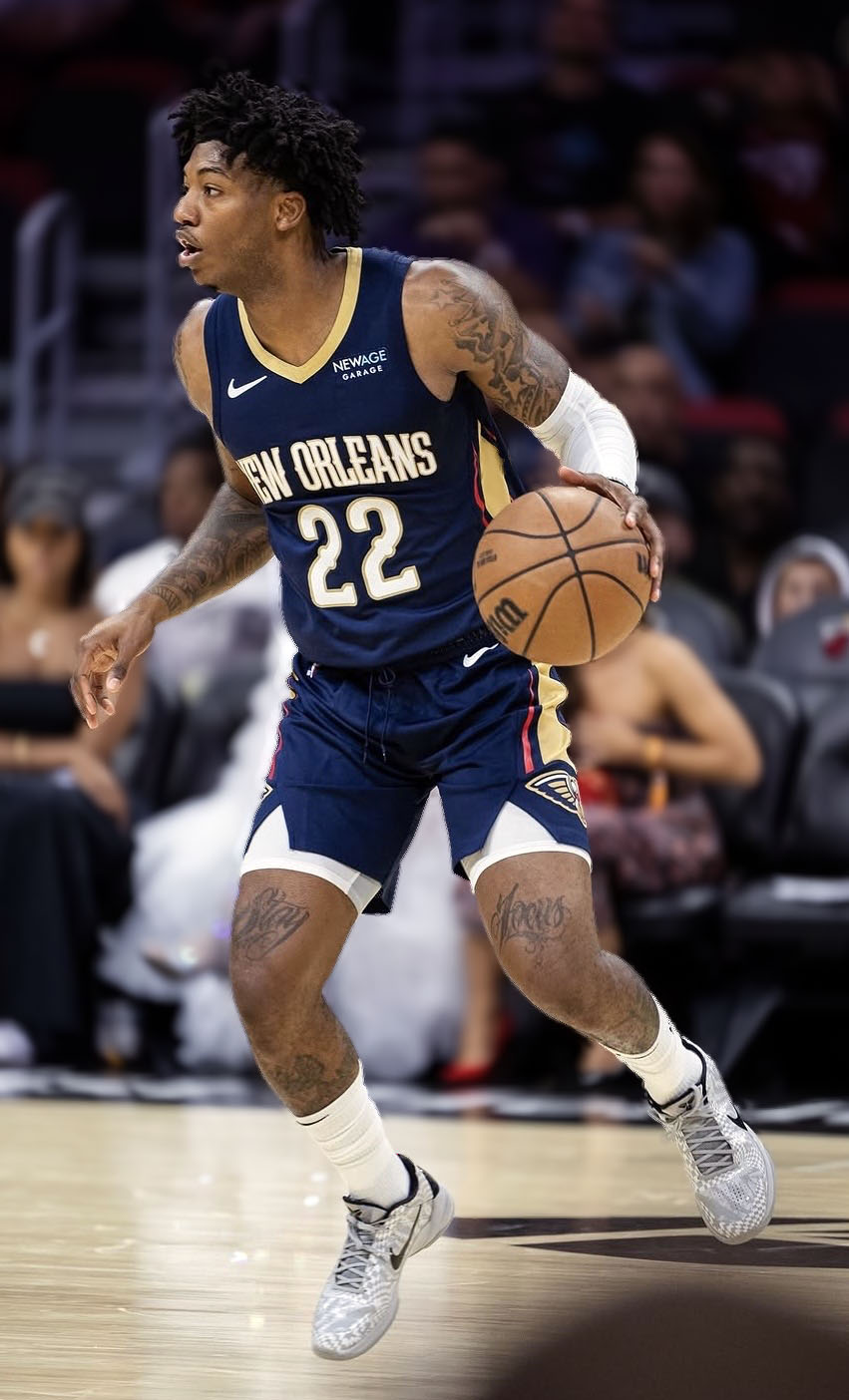 Elfrid Payton in a navy New Orleans Pelicans uniform dribbles a basketball on the court during a game. Spectators are visible in the blurred background.