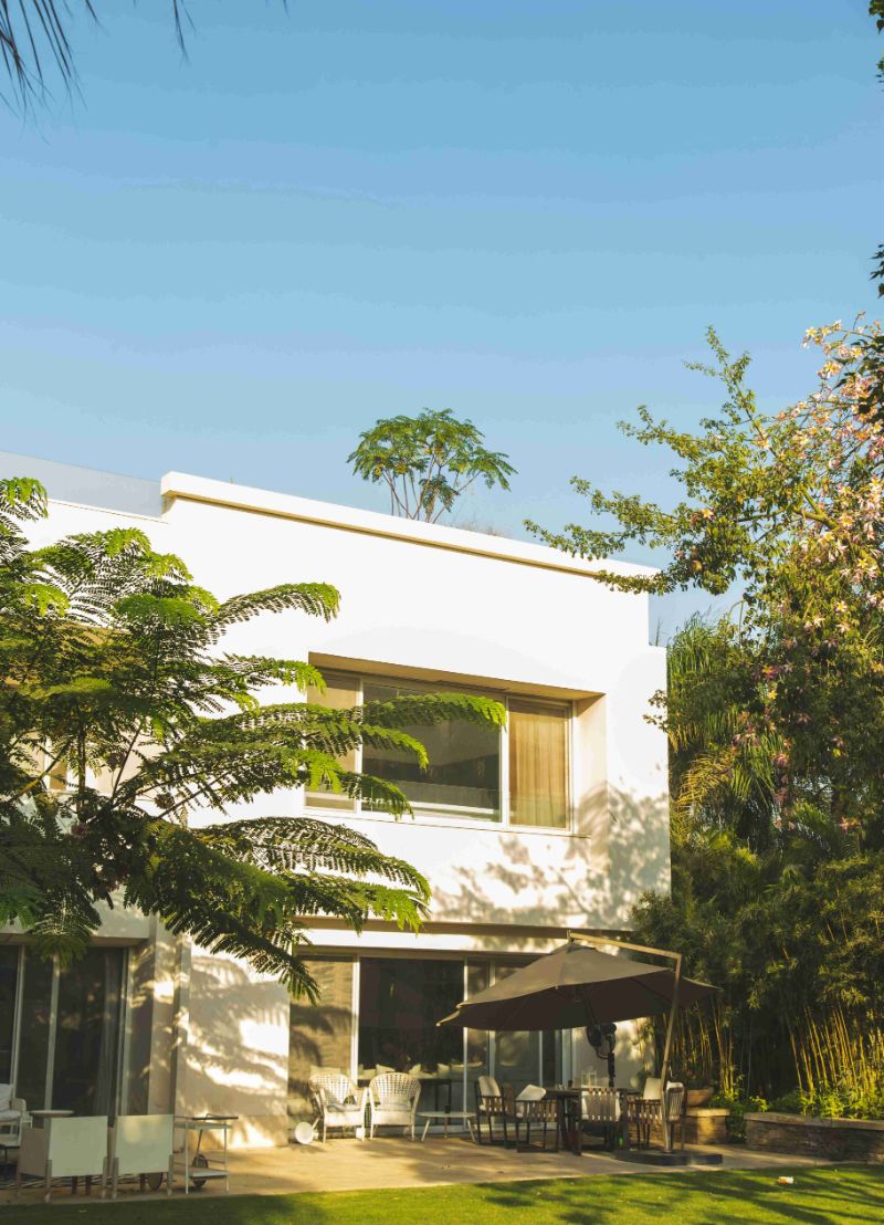 Modern white villa with a sunny patio, brown umbrella, and chairs surrounded by lush tropical trees and a green lawn.