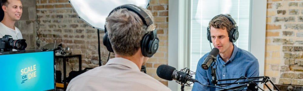 man in gray shirt leaning on table with headphones facing another man leaning on table with headboard