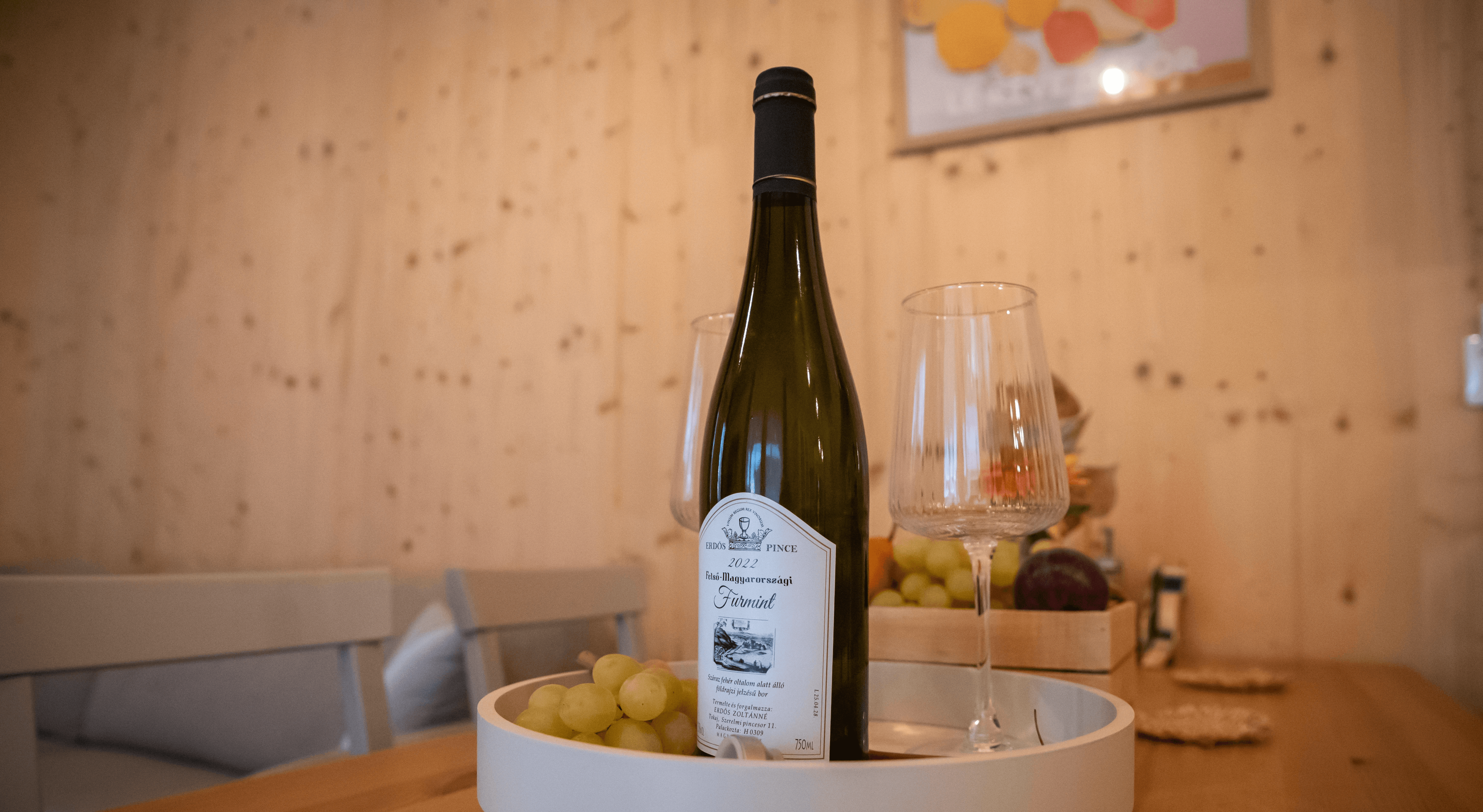 Egy palack fehérbor elegánsan van elrendezve két borospohárral és egy tányér zöld szőlővel egy faasztalon, világos fa fal előtt, meghitt étkezési hangulatot teremtve.