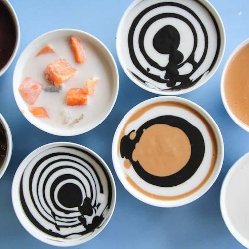 Several bowls of desserts on a blue surface, some with black and white circular patterns and one with orange chunks in white liquid.