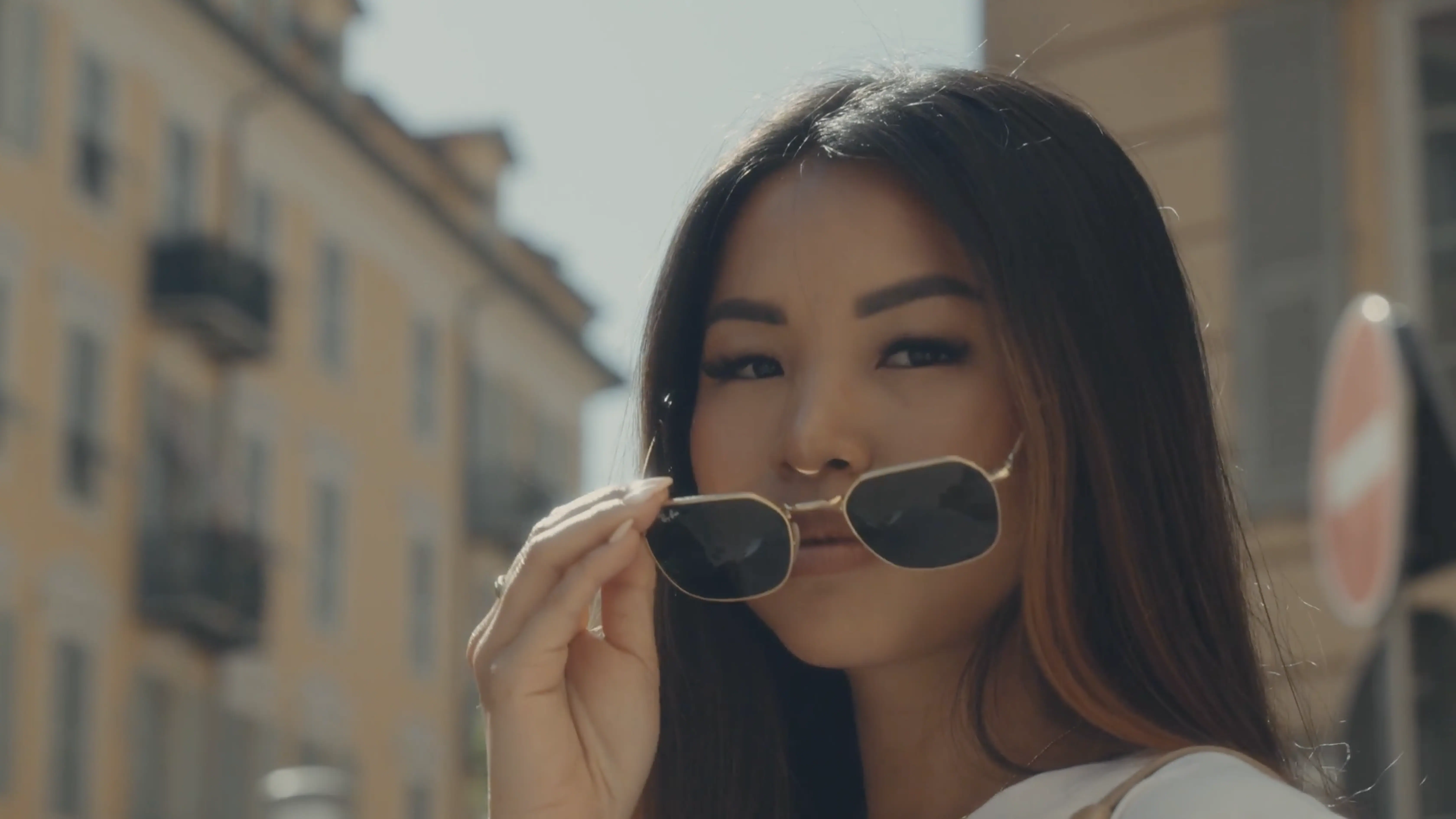 A person with long hair holds sunglasses to their face while standing on a sunny street, with buildings and a traffic sign in the background.