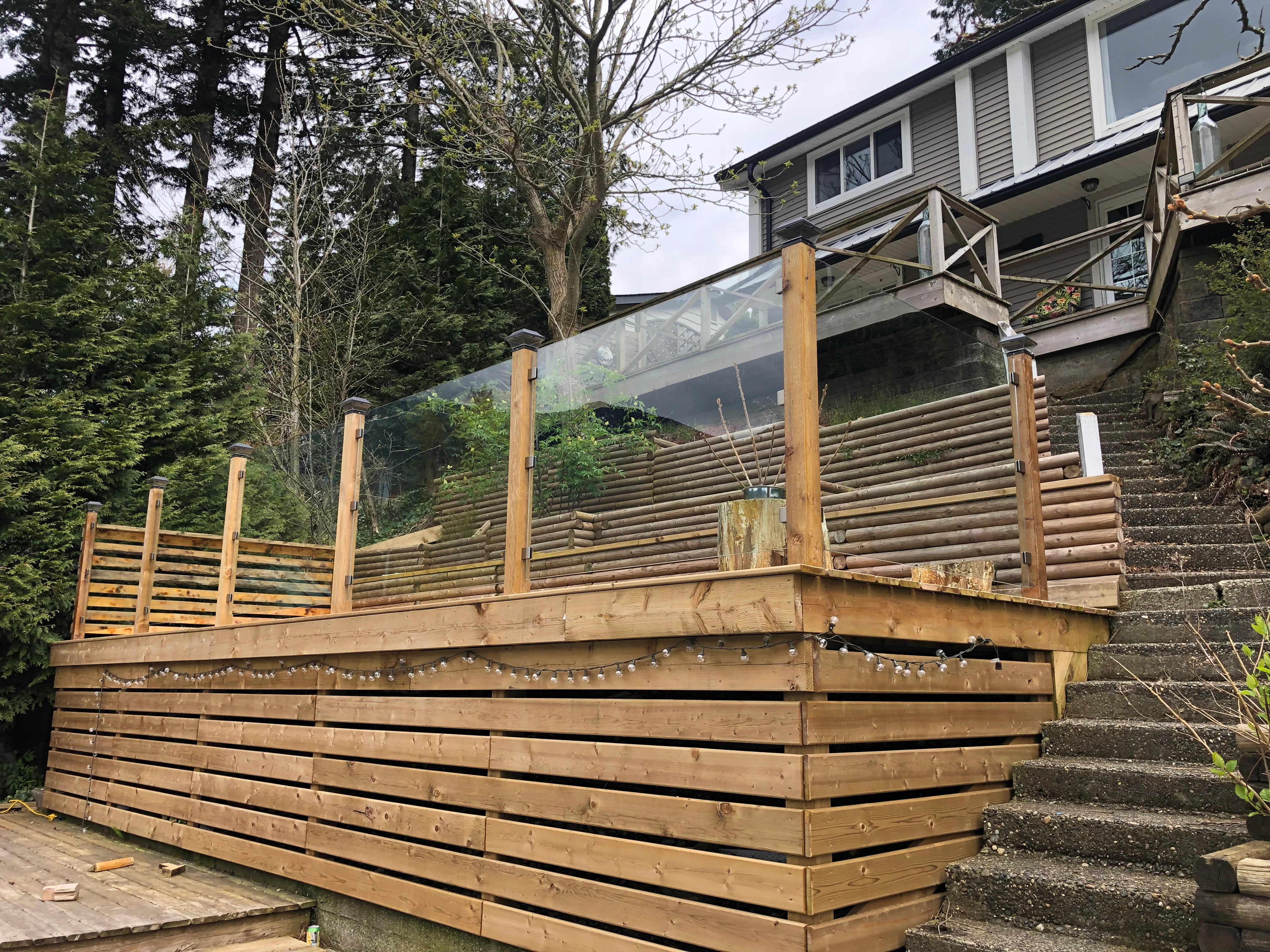 A rustic outdoor wooden deck on a sloped hillside property featuring clear glass panels installed between thick wood posts.