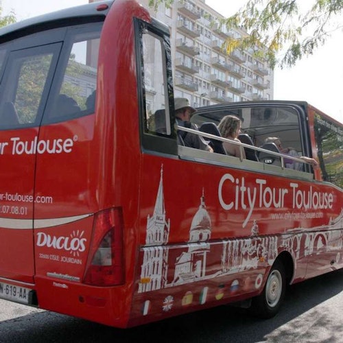 Ônibus de CityTour Vermelho com passageiros, teto aberto, e marcos de Toulouse ilustrados nas laterais, dirigindo próximo a um edifício residencial.