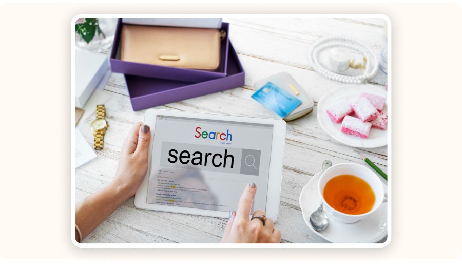 Someone using a tablet at a cozy table surrounded by everyday items — credit card, purse, tea, and snacks — while manually searching for SEO competitors.