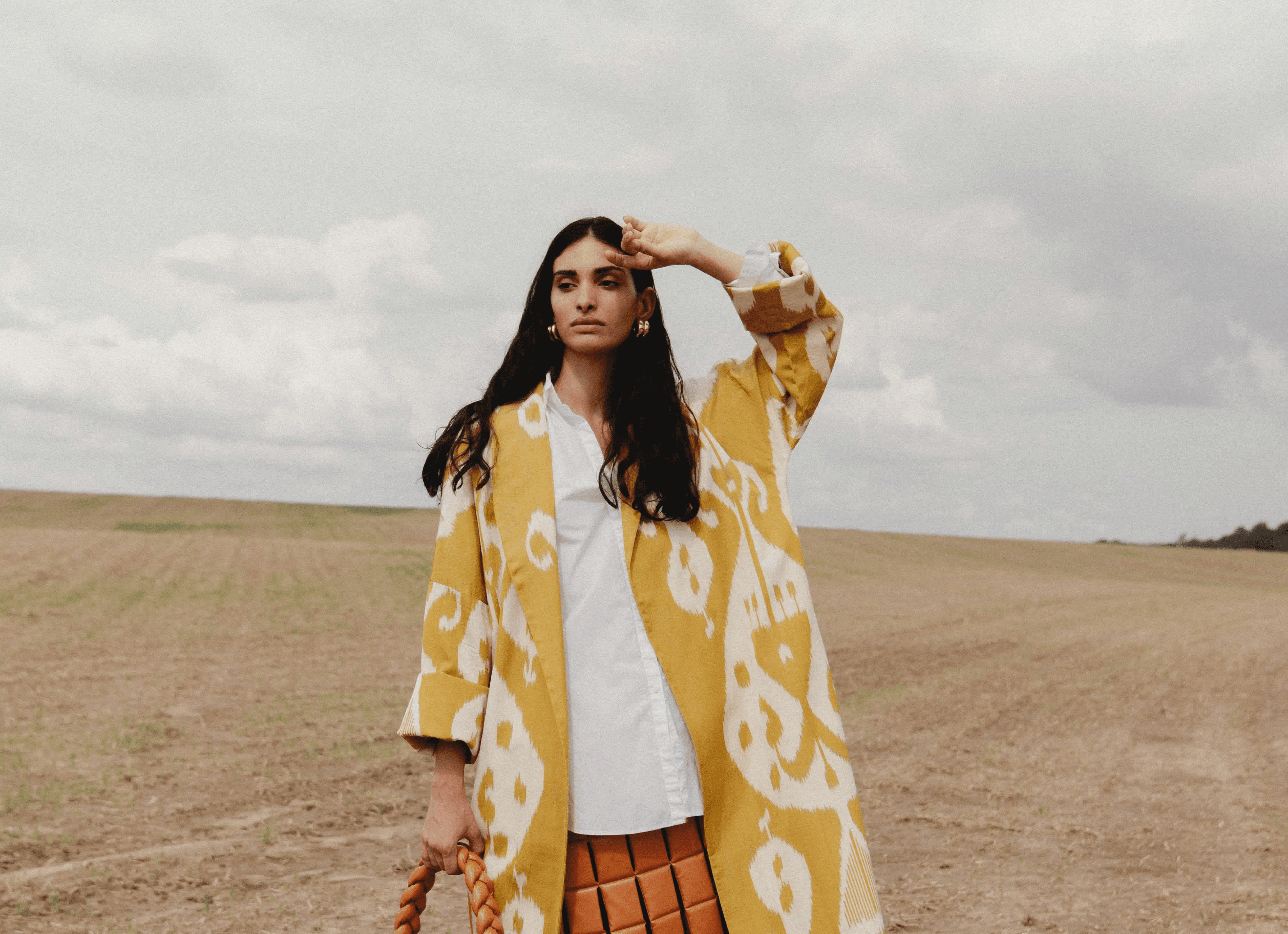 Woman standing in a field wearing a white shirt and long yellow patterned coat, shielding her eyes from the sun.
