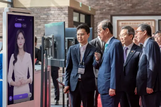 บุคคลสำคัญรวมถึงนายกรัฐมนตรีดัก-ซู ฮัน สังเกตบริการ AI Human ของ ESTsoft ซึ่งทำงานบนจอคีออสแนวตั้ง