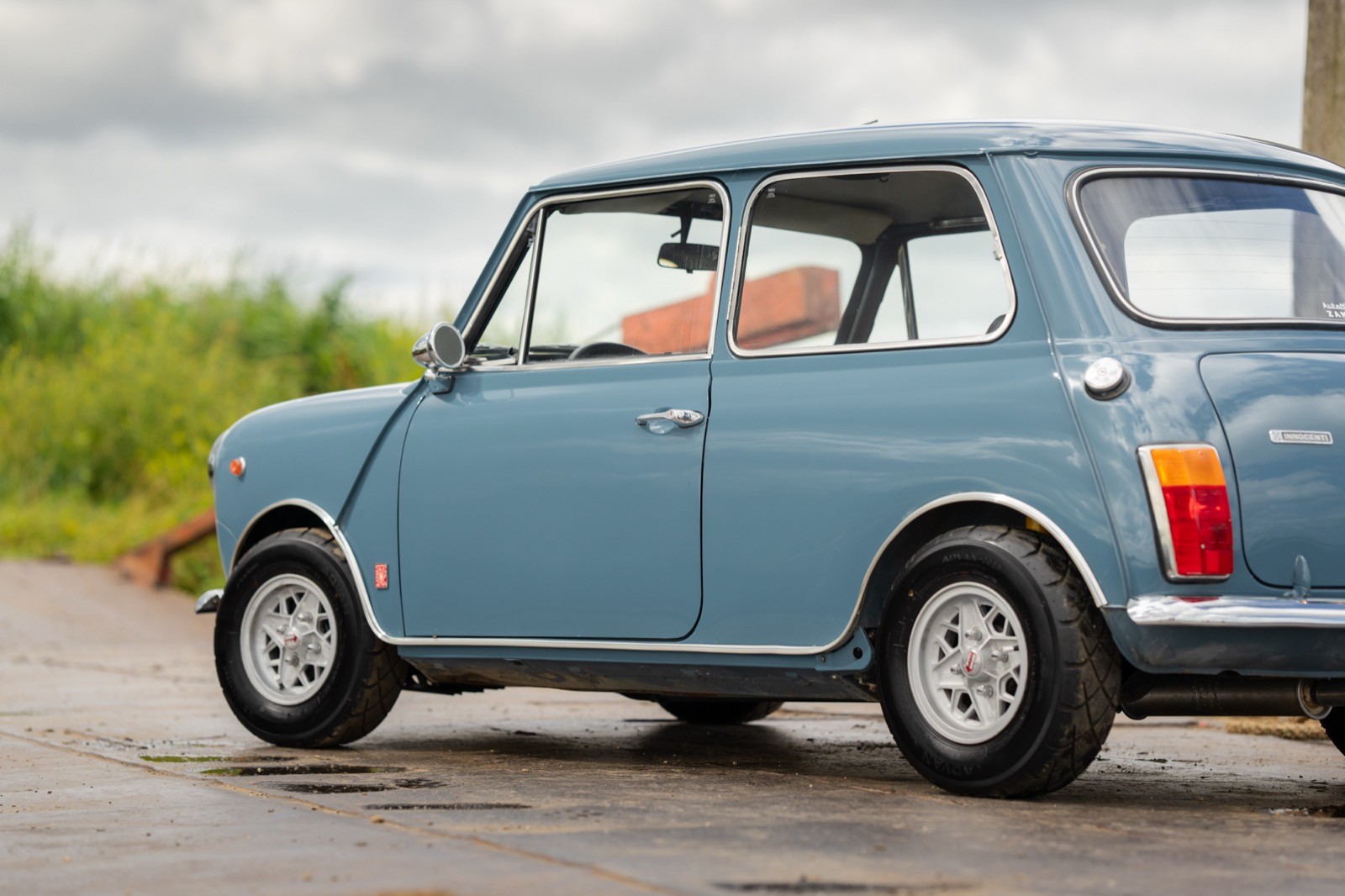 A classic blue car parked on a road with green scenery in the background, partially showing the driver inside.