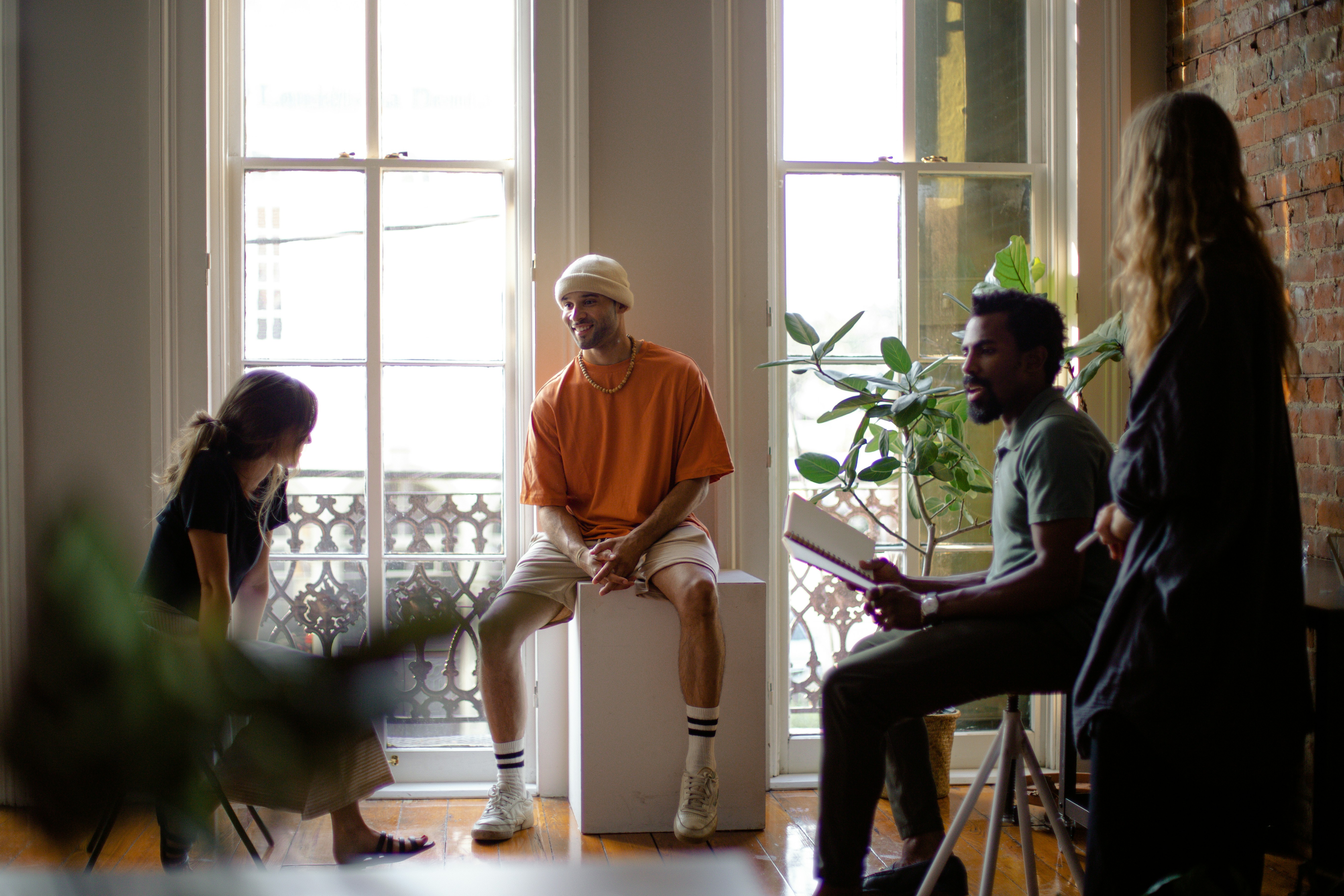A group of four people engaged in discussion in a bright, minimalist room with large windows and plants.