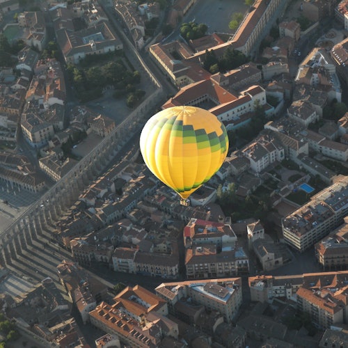 A yellow and green hot air balloon floats over a densely packed town with orange-tiled roofs and narrow streets.