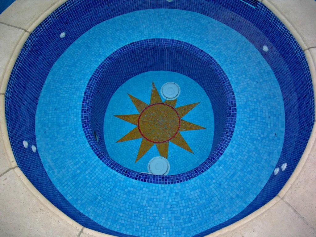 Hand-cut sun medallion set into the floor of a round spa, seen through the water — Mosaiclegs Art Studio