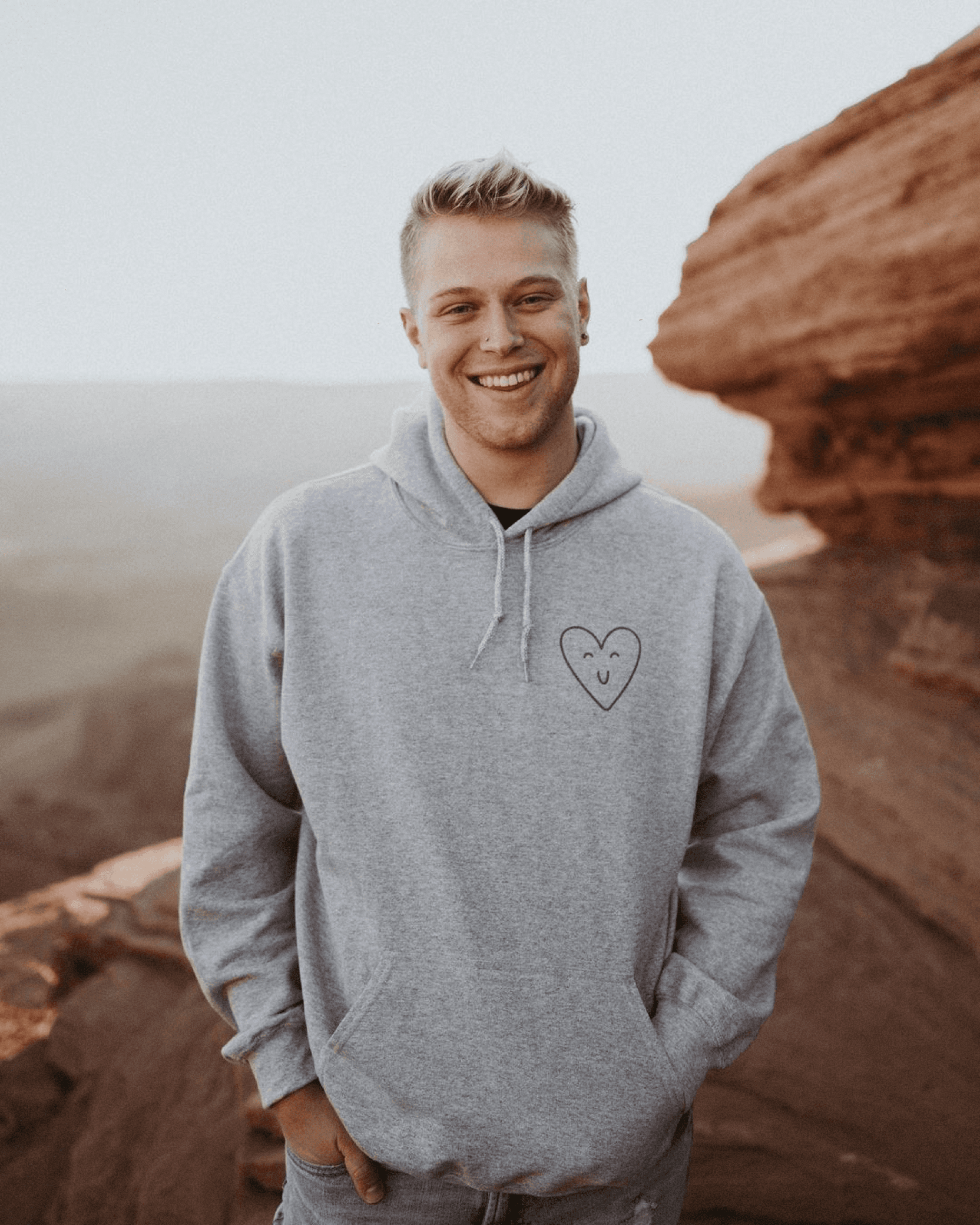 Smiling man in gray hoodie with small heart design, standing outdoors against a rocky landscape.
