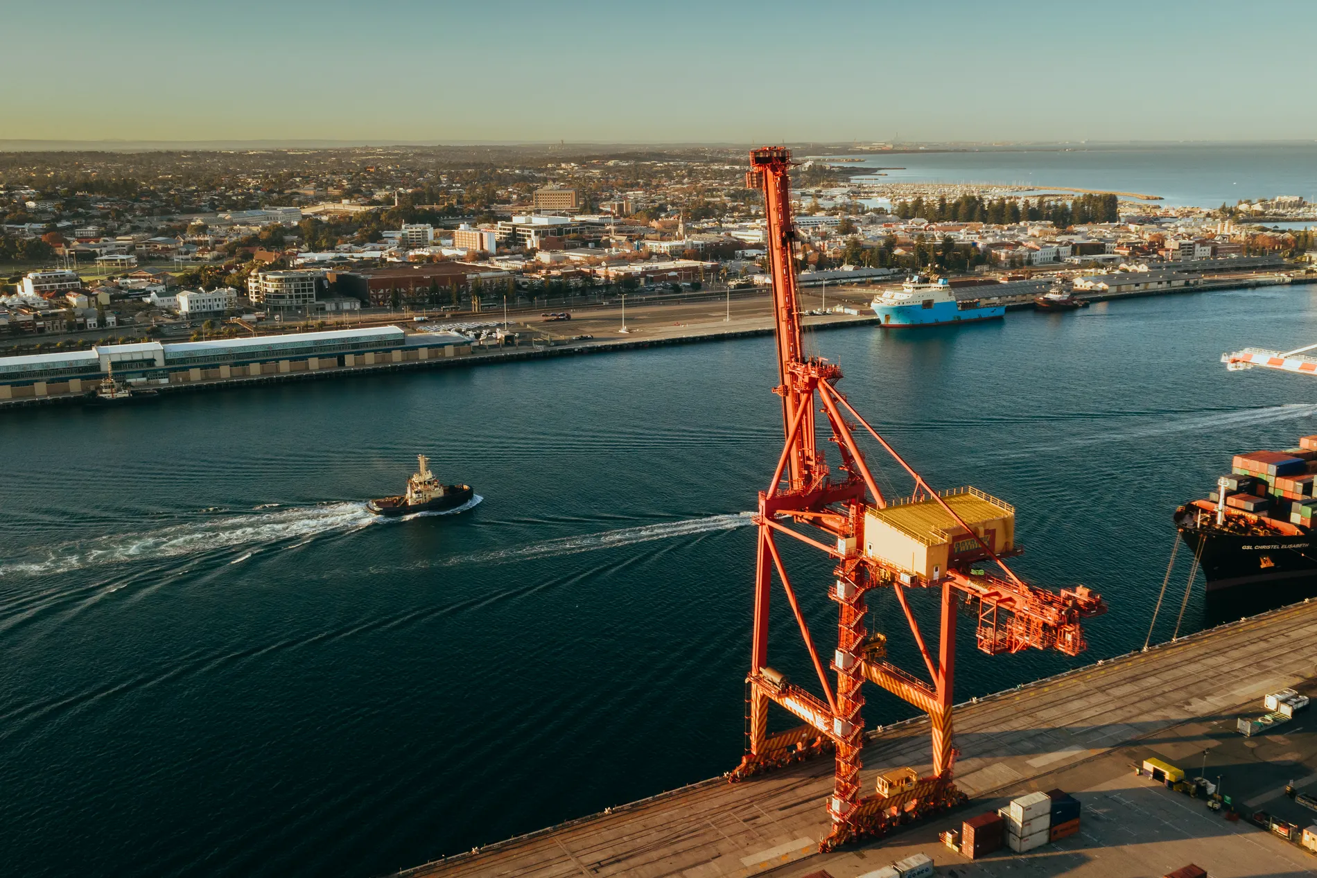 A red harbour crane beside a channel at golden hour, a tug under way and a town beyond