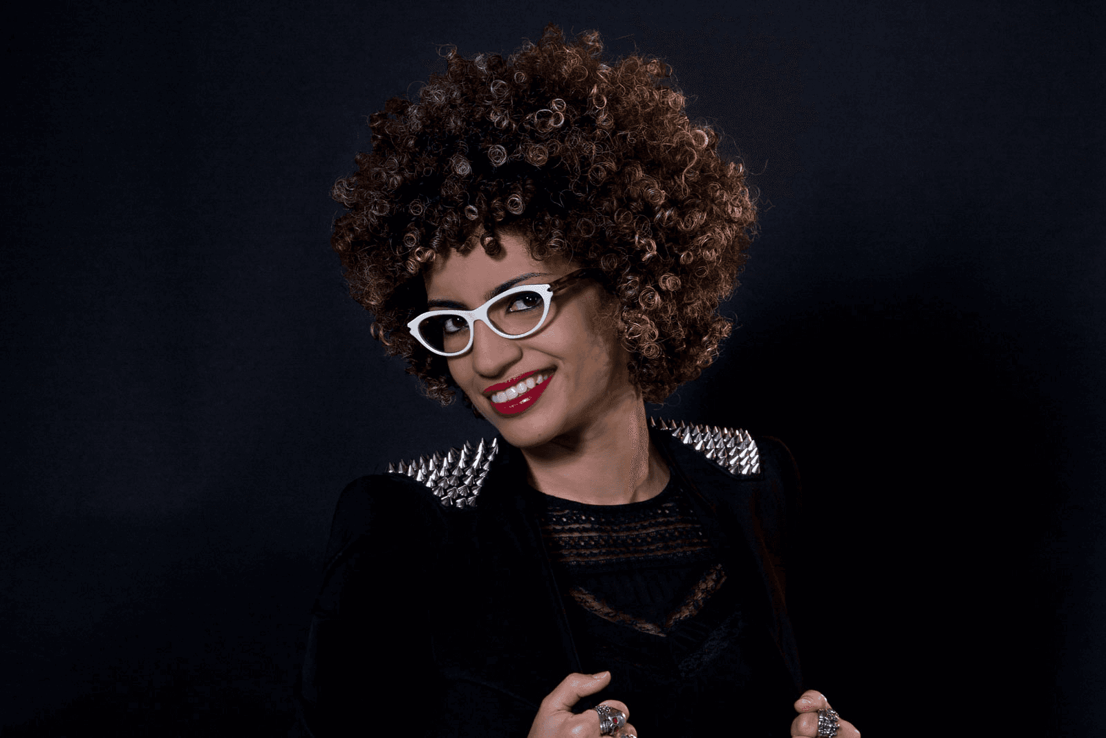 A woman with curly brown hair wears white-framed glasses and a black top with silver stud details. She is smiling and looking directly at the camera against a dark background. Her hands are clasped in front of her.
