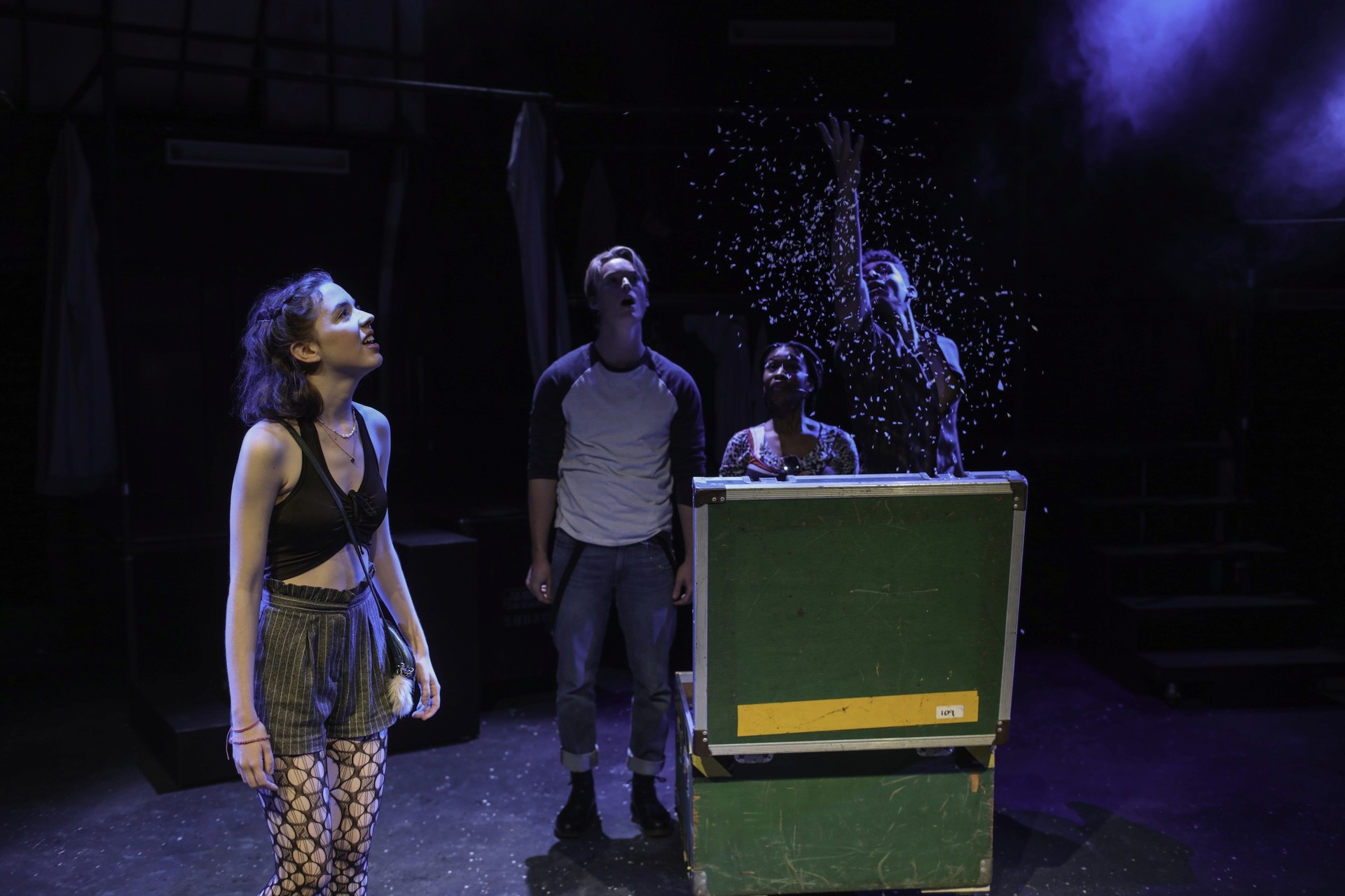Four performers gather around a large green road case as artificial snow bursts into the air. One performer stands downstage watching in wonder, illuminated by cool stage lighting.