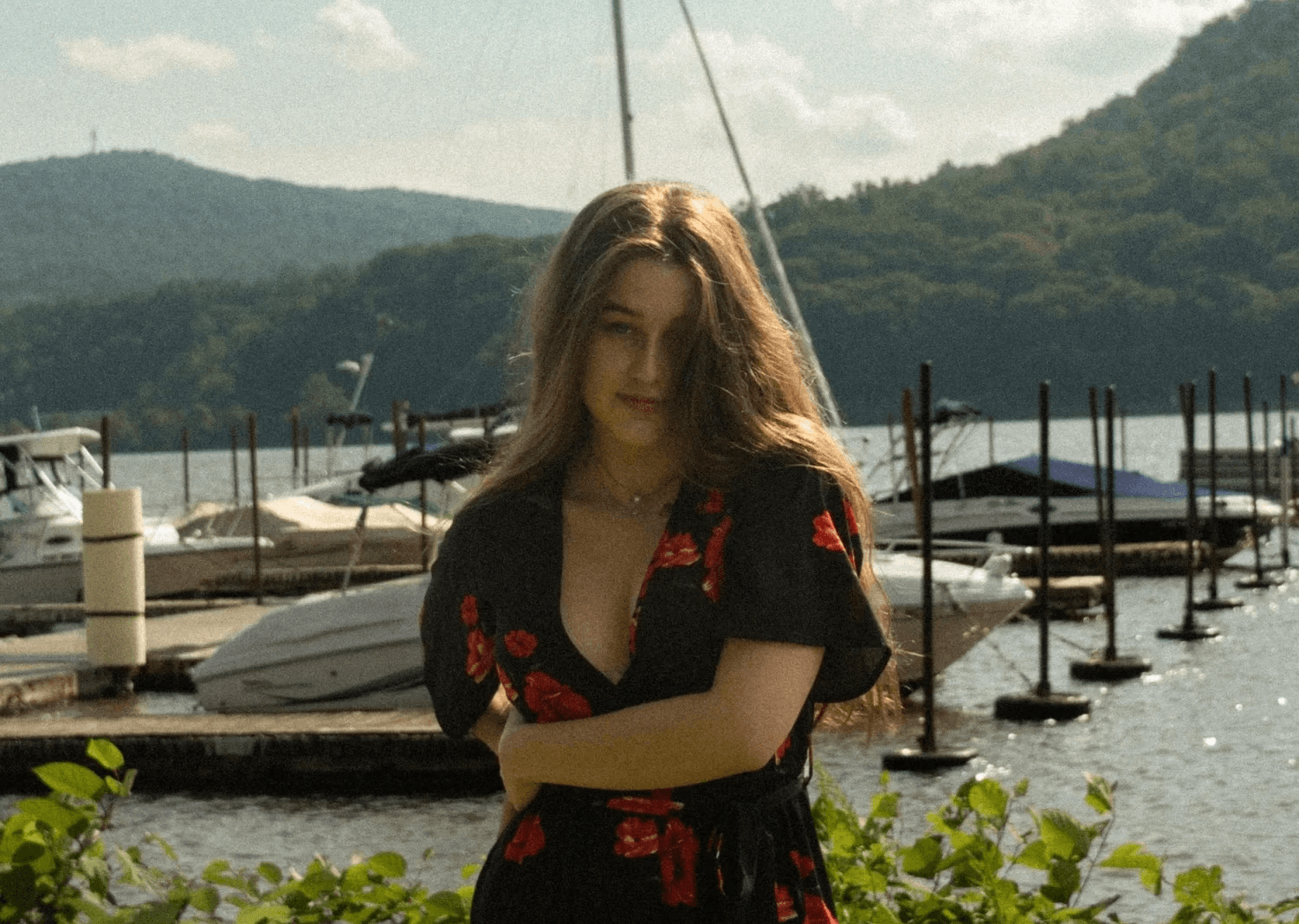 Varvara Aristakesyan stands at a marina wearing a black floral dress with red roses, arms crossed, looking directly at the camera with a calm expression. Sailboats, a dock, and green hills are visible in the background.