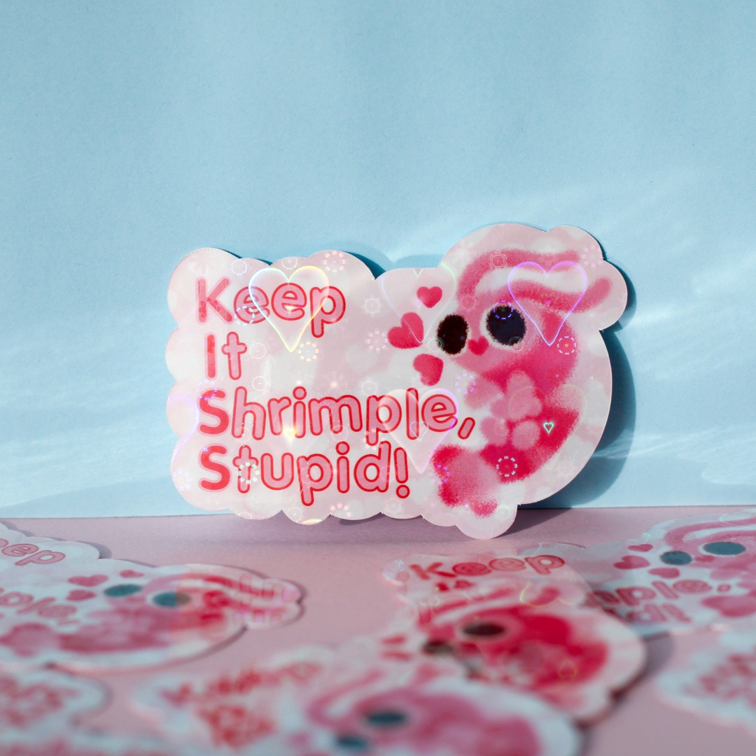 A light pink sticker that reads "Keep it Shrimple, Stupid!" with an ilustration of a cute shrimp. It has a holographic laminate that reflects heart shapes.