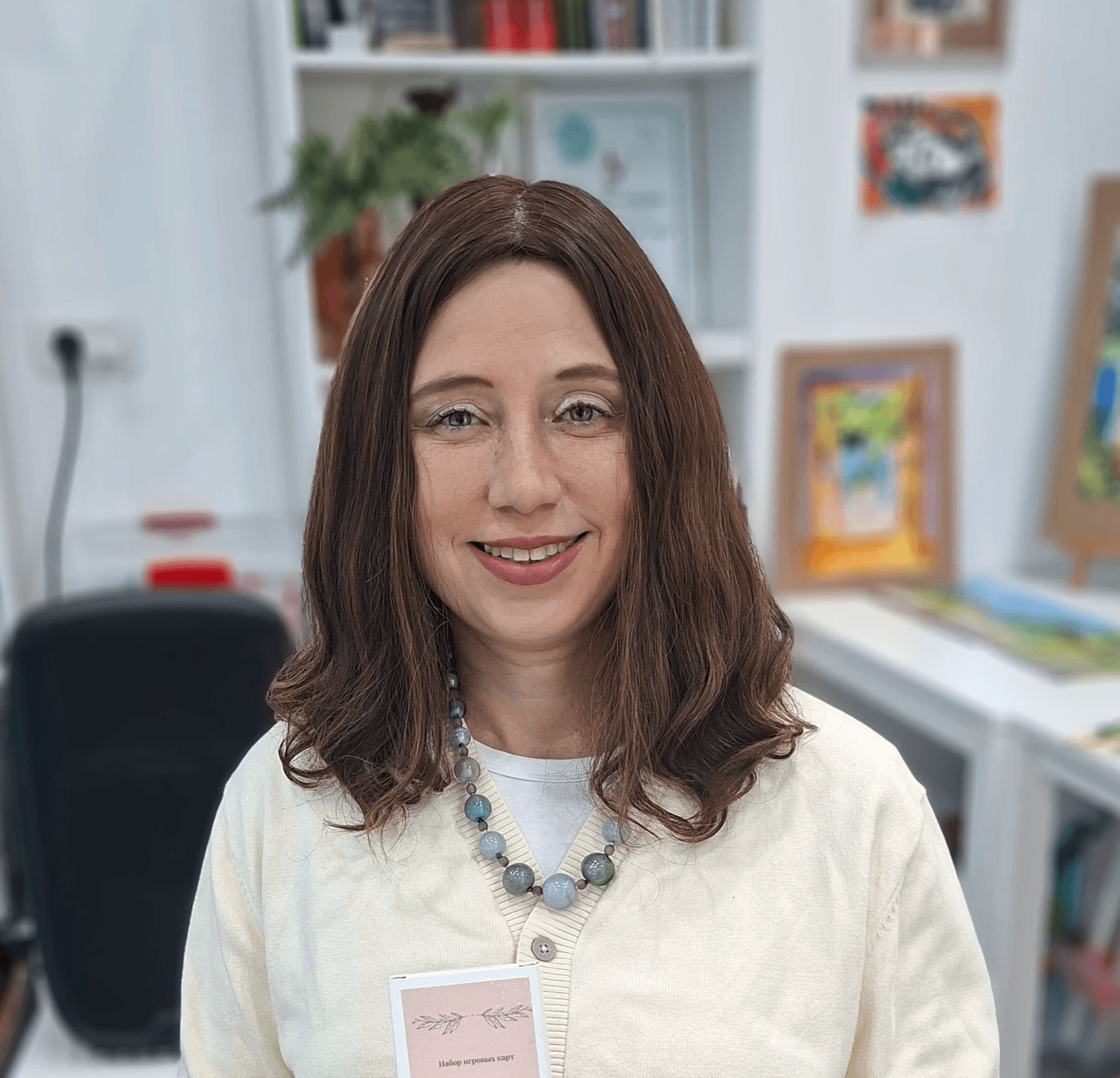 Smiling female therapist with short gray hair, wearing a brown top and a purple shawl, surrounded by a cozy background.