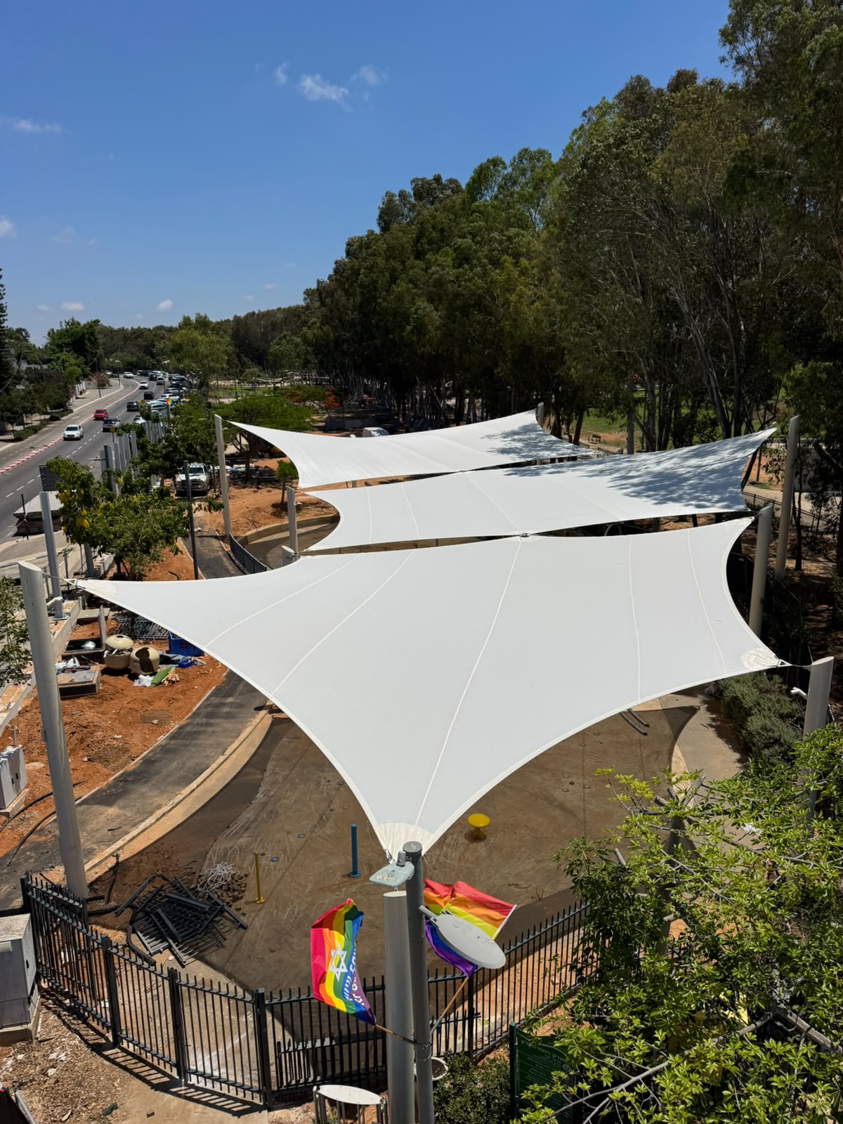 Three white, sail-shaped sunshades over a walkway next to a road with trees in the background.