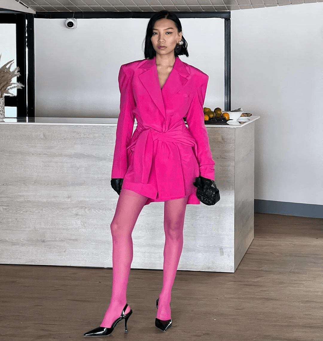 Ikvison Jamang sitting on a red stool in front of a bright red ad for Diesel. She is wearing retro jeans and a sheer top with shades and a white handbag.  