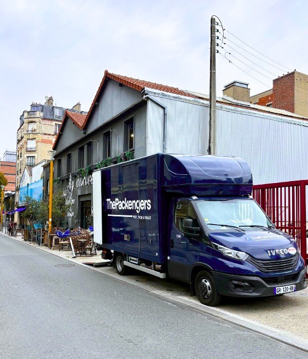 A blue ThePackengers transport truck parked on a narrow European city street, highlighting premium urban collection and delivery services for high-end furniture.