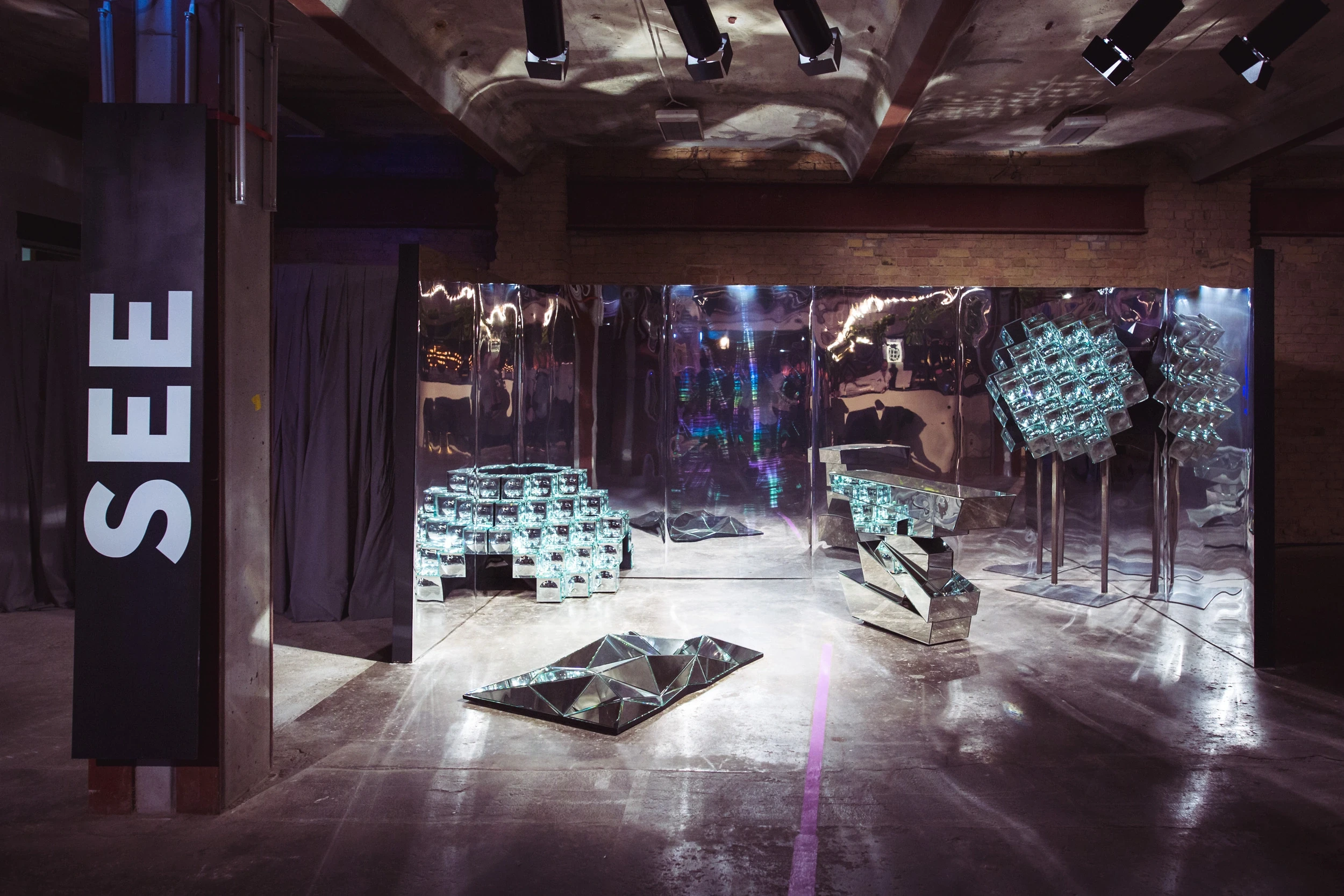 Mirror installation at DIAGEO BCB18 with faceted reflective sculptures, mirrored walls and a SEE sign on a wooden pillar.