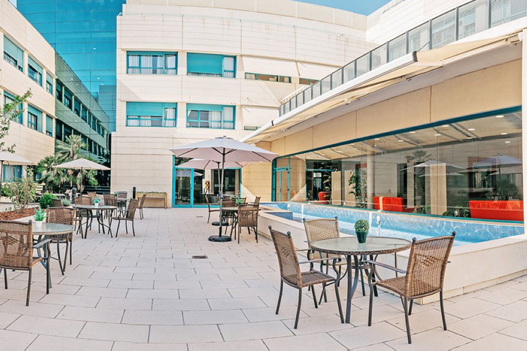 Outdoor terrace and patio area with dining tables and chairs, colorful umbrellas, bright blue sky, building facade, and landscaped surroundings