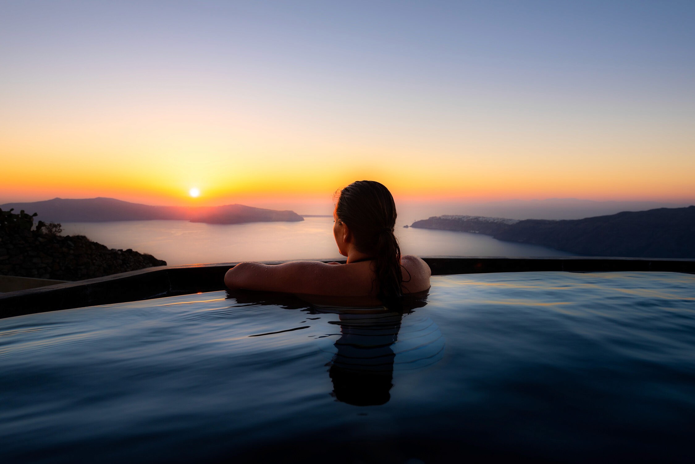 A person relaxes in an infinity pool at sunset overlooking a misty mountain range and valley.