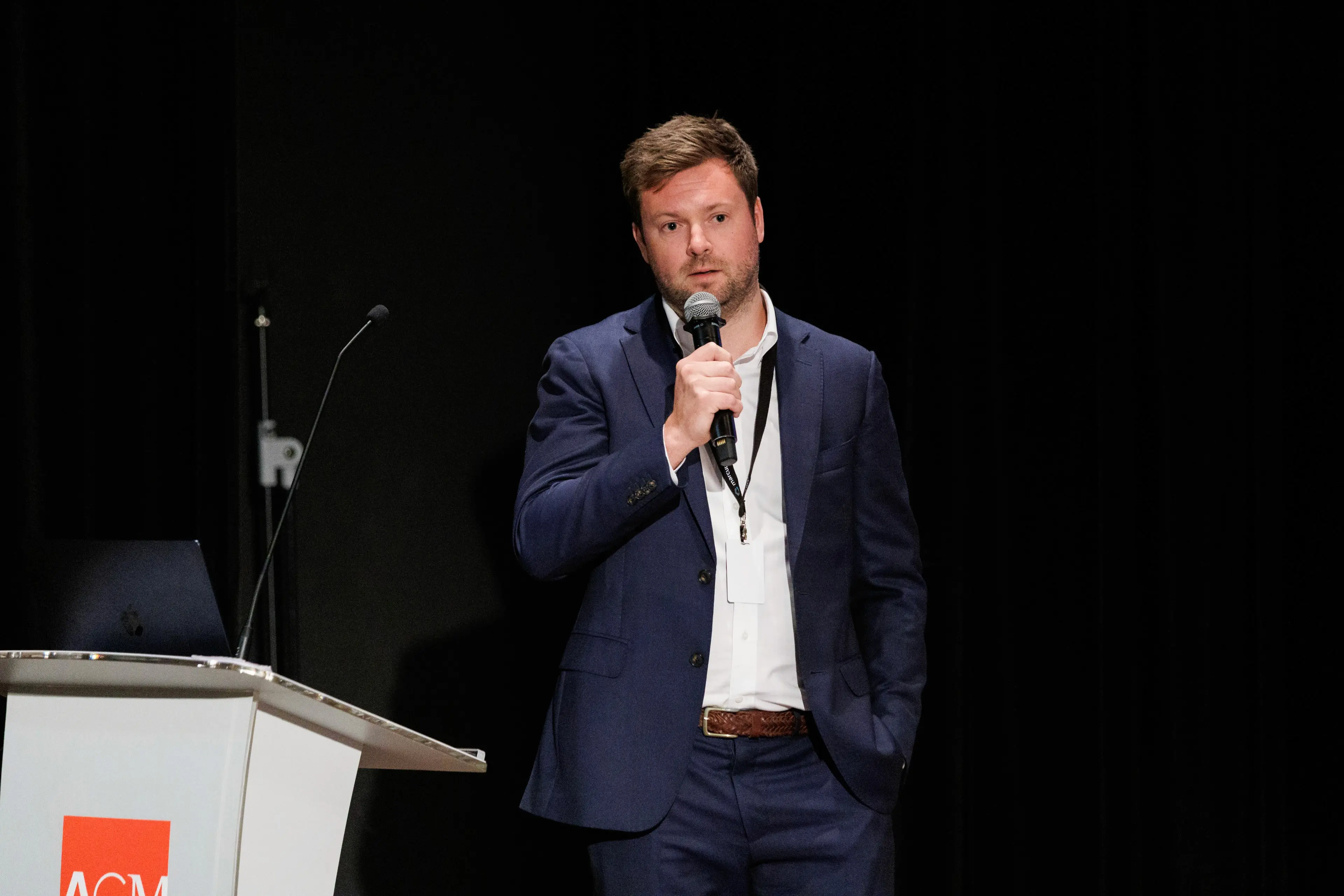 A man in a suit speaks into a microphone at a podium during an event, with a focused expression.